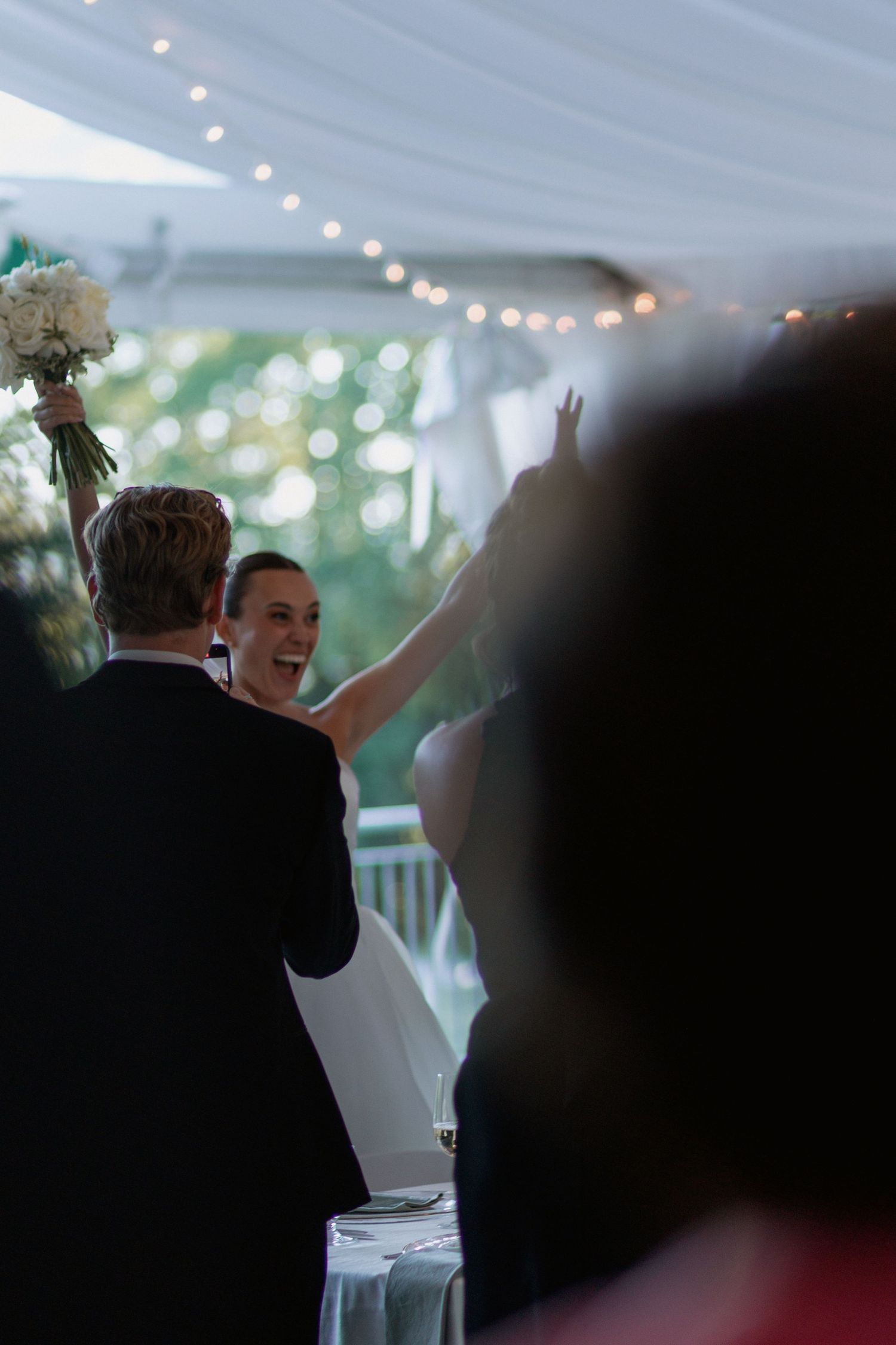 Celebratory moment at a wedding reception with guests raising their arms under twinkling lights.