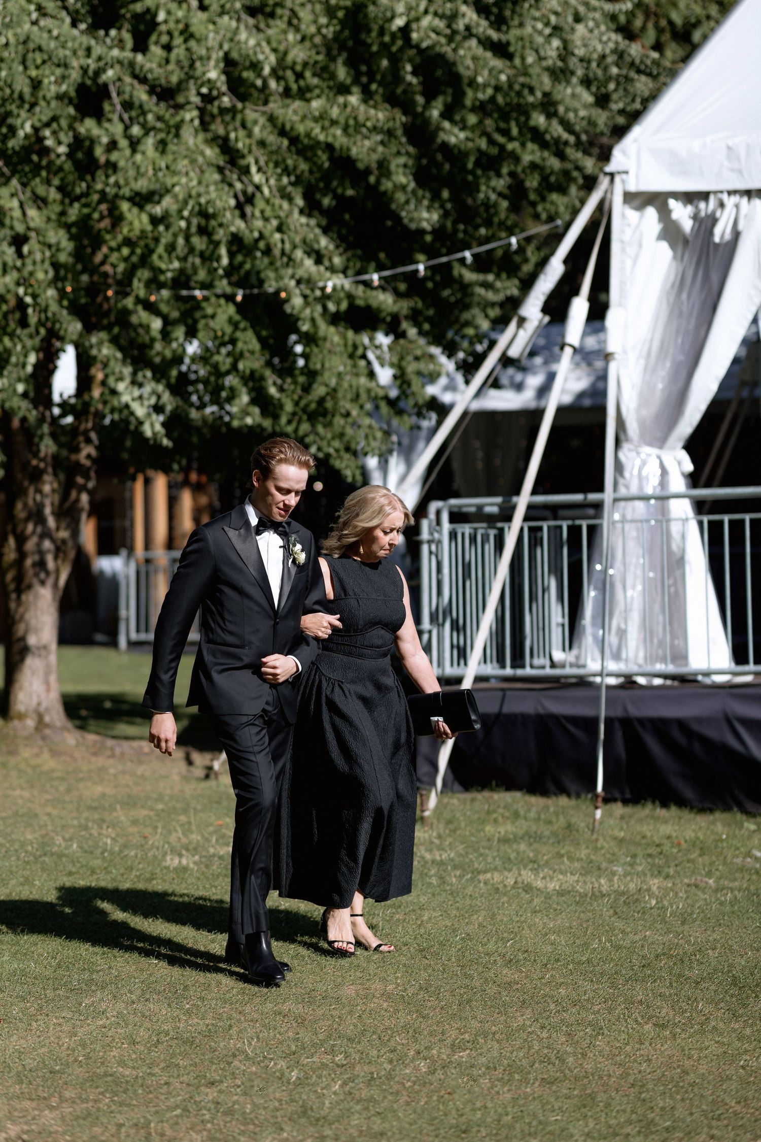 Wedding attendees in formal black attire walking near white reception tent.