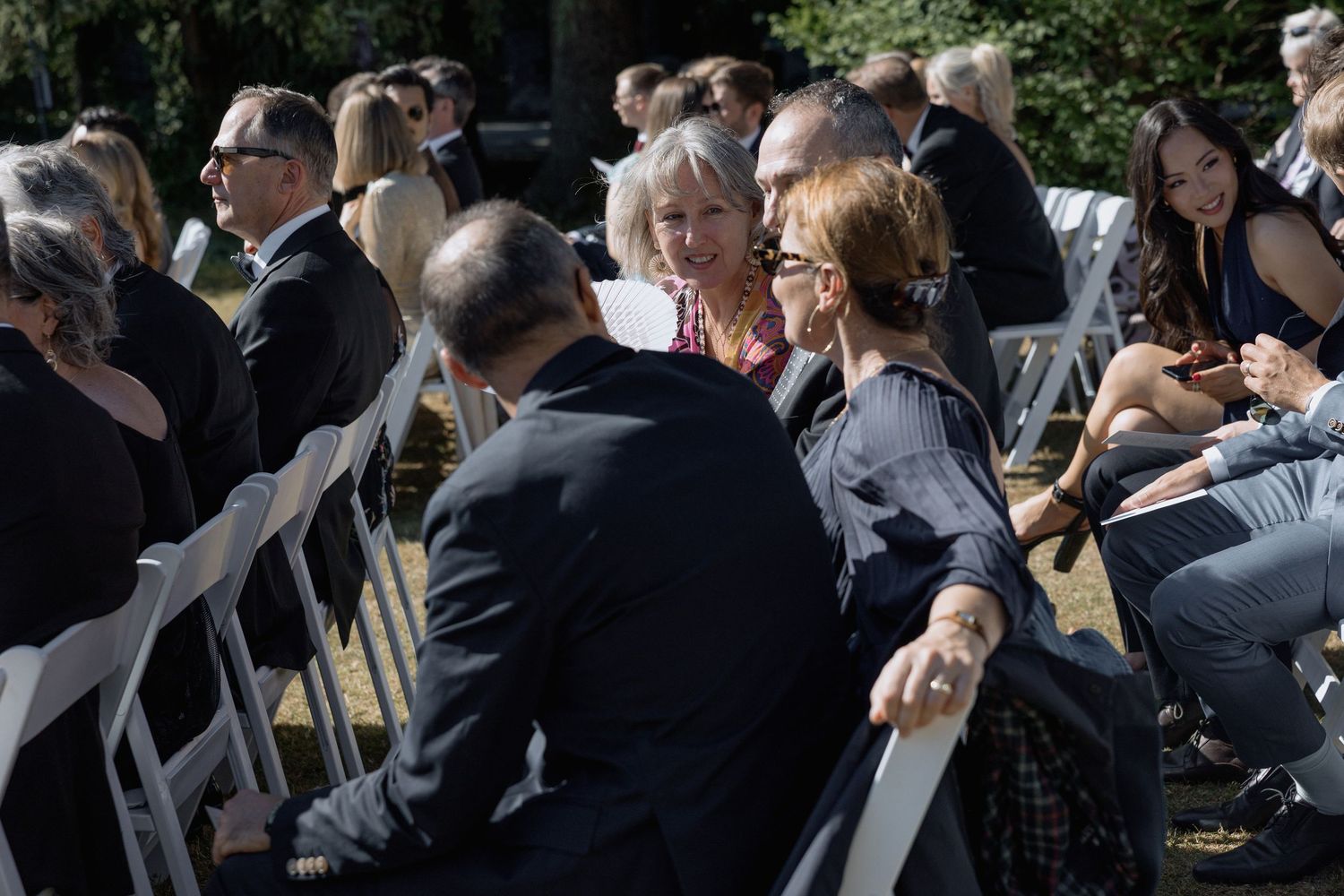 Wedding attendees seated in white chairs watch the ceremony proceedings in a shaded outdoor setting.