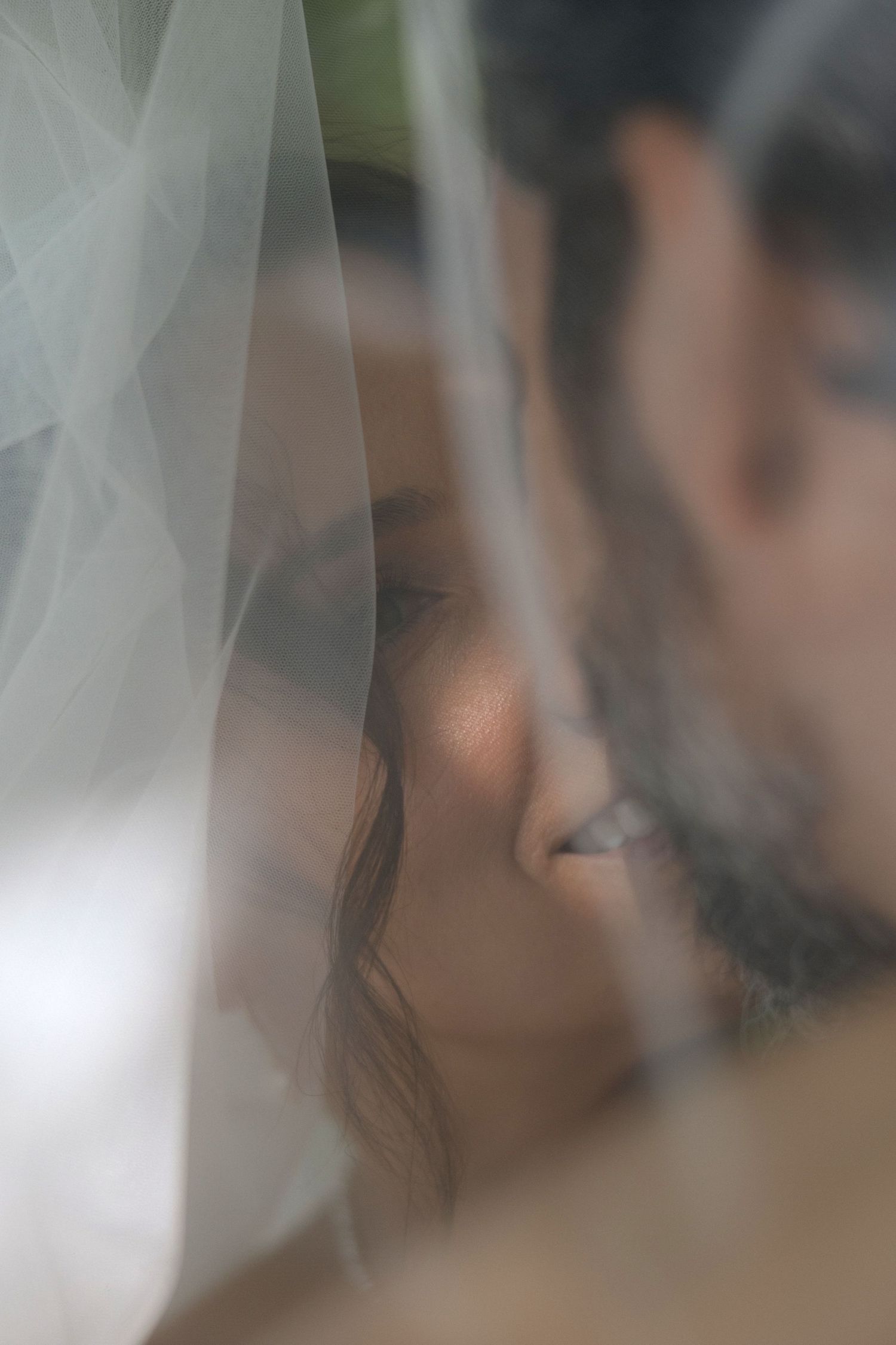 A soft focus artistic wedding photo showing a delicate white veil with blurred figures behind it.