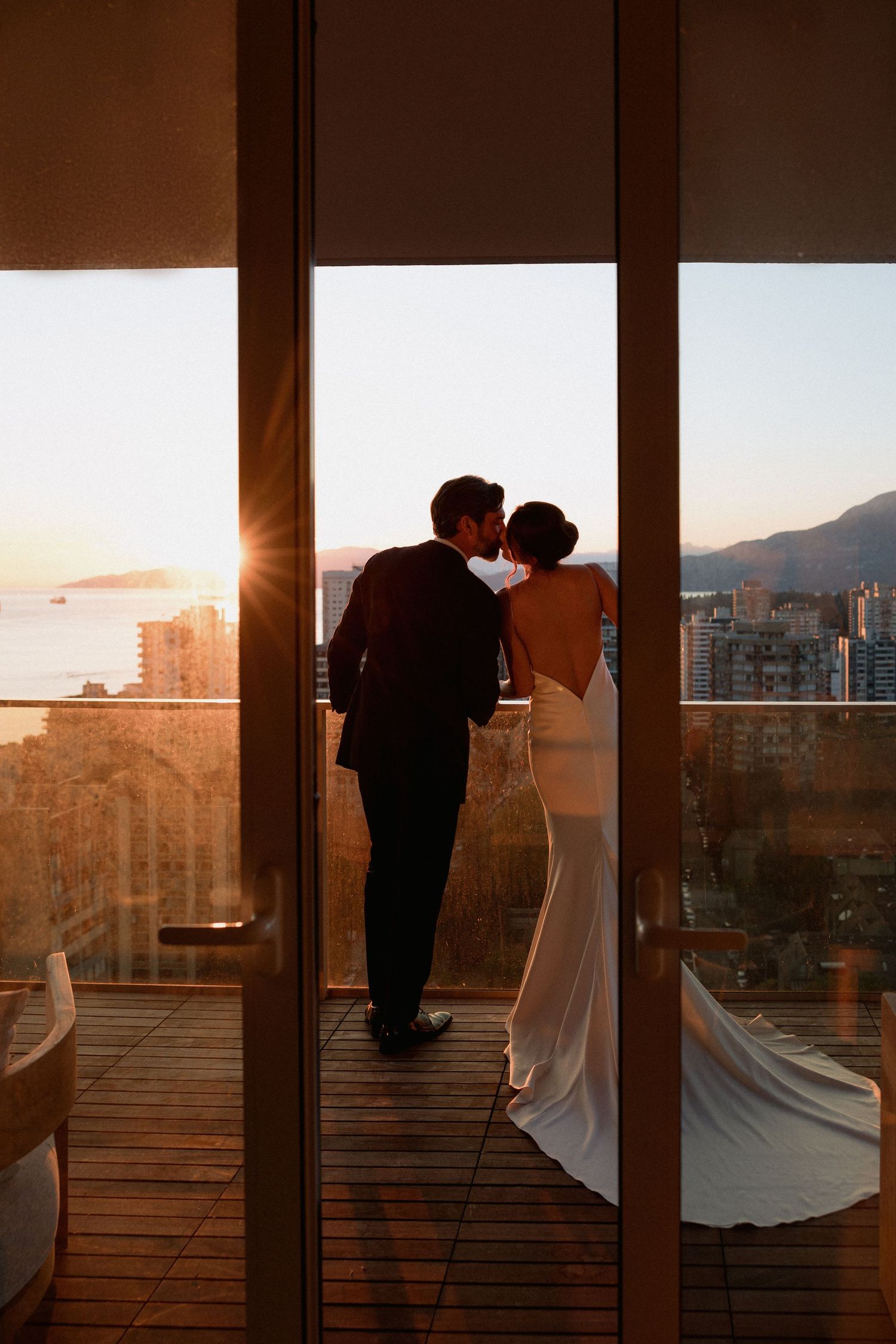 Silhouetted newlyweds share an intimate moment against floor-to-ceiling windows with city views at dusk.