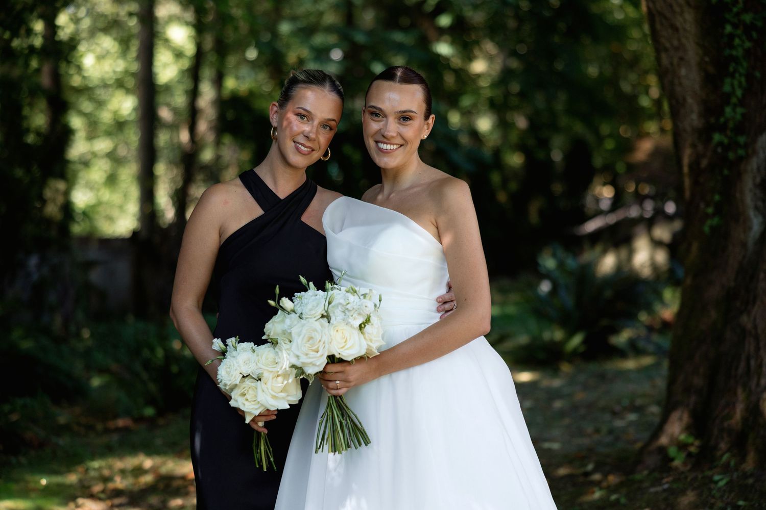 Candid wedding photo showing two people smiling with white floral bouquets in a garden setting.