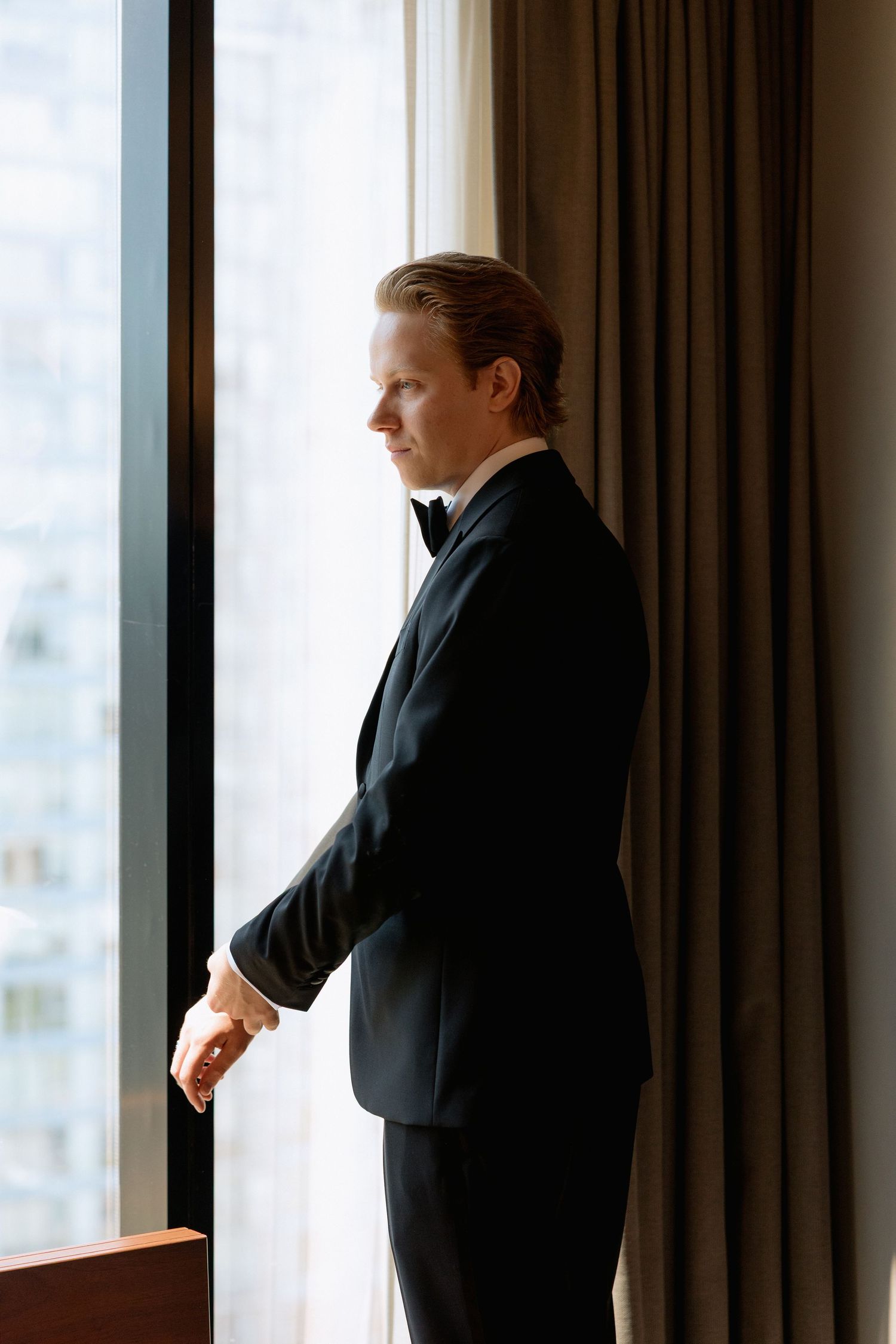 Groom in dark suit standing contemplatively by large hotel window at Marriott Hotel in Vancouver