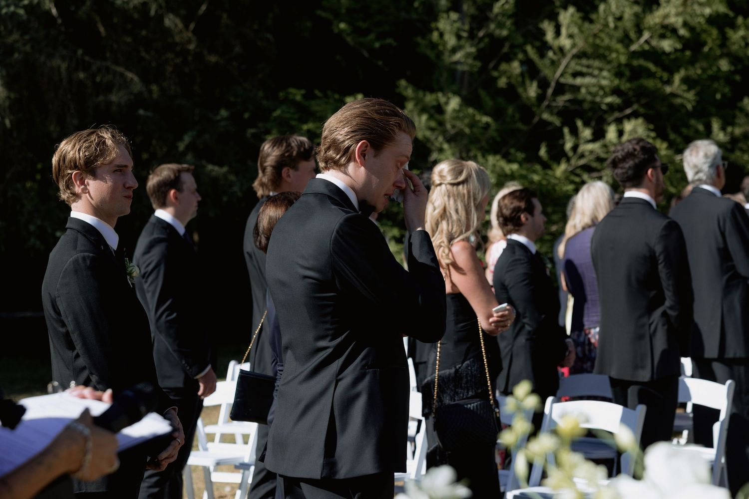Wedding guests standing during an outdoor ceremony with white chairs arranged in rows.