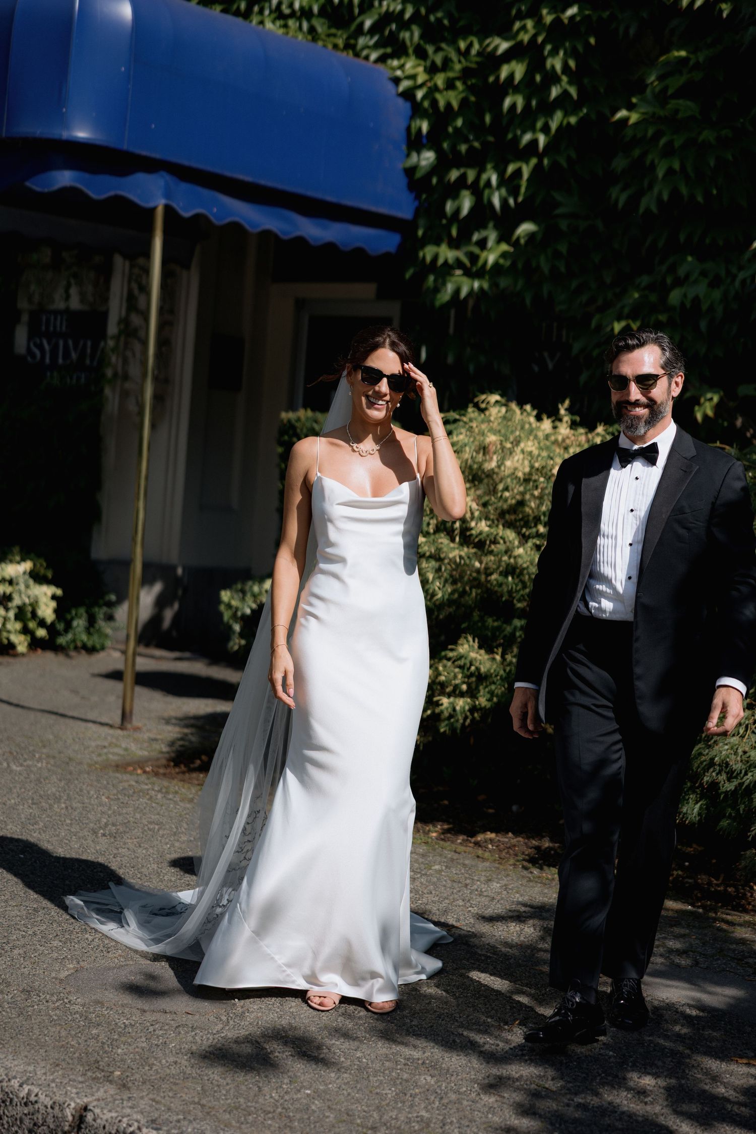Elegant couple in formal wedding attire walking outdoors with a blue awning in the background in front of the Sylvia Hotel in Vancouver