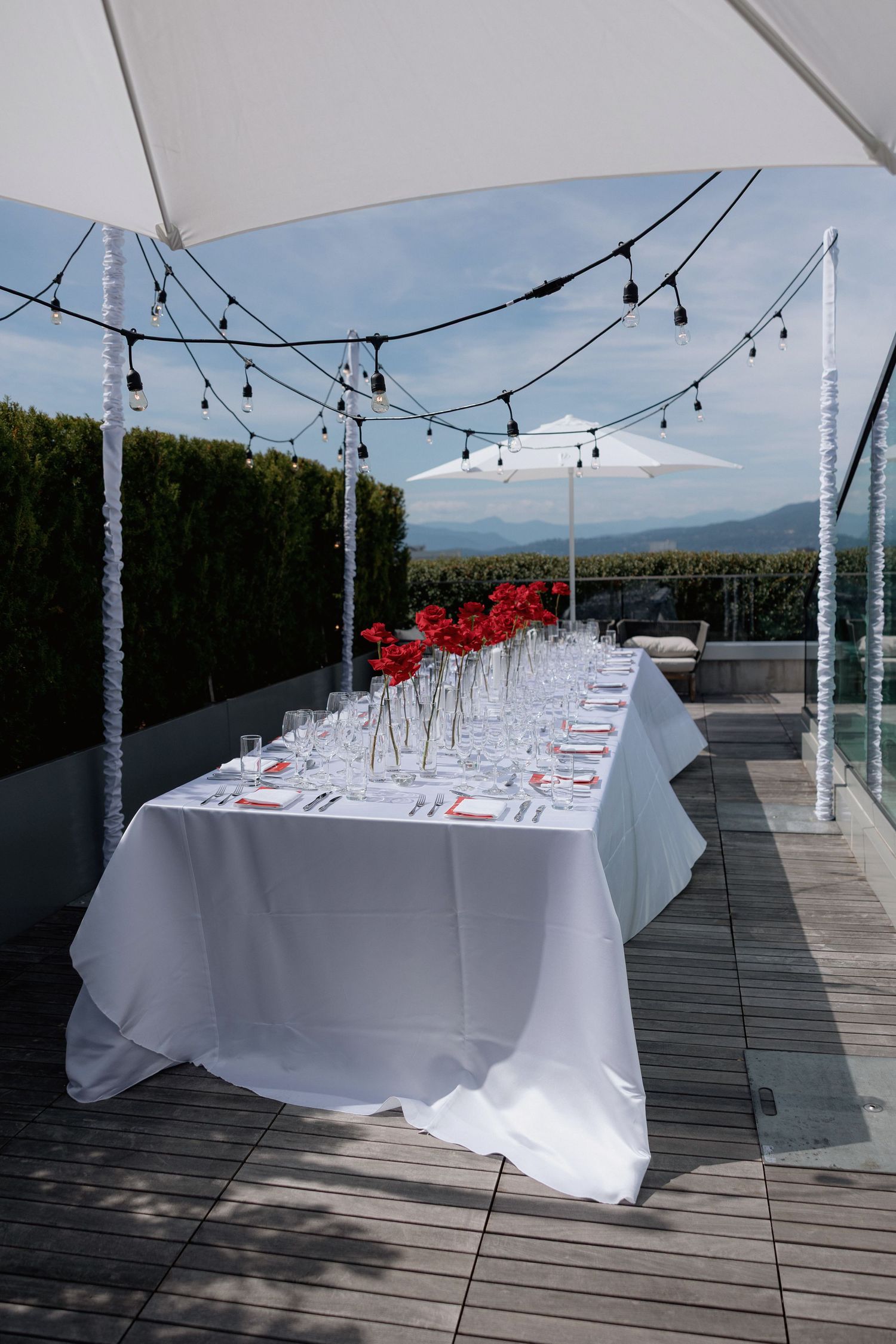 An elegant outdoor dining setup with a long white-clothed table and string lights overlooking a mountain landscape.