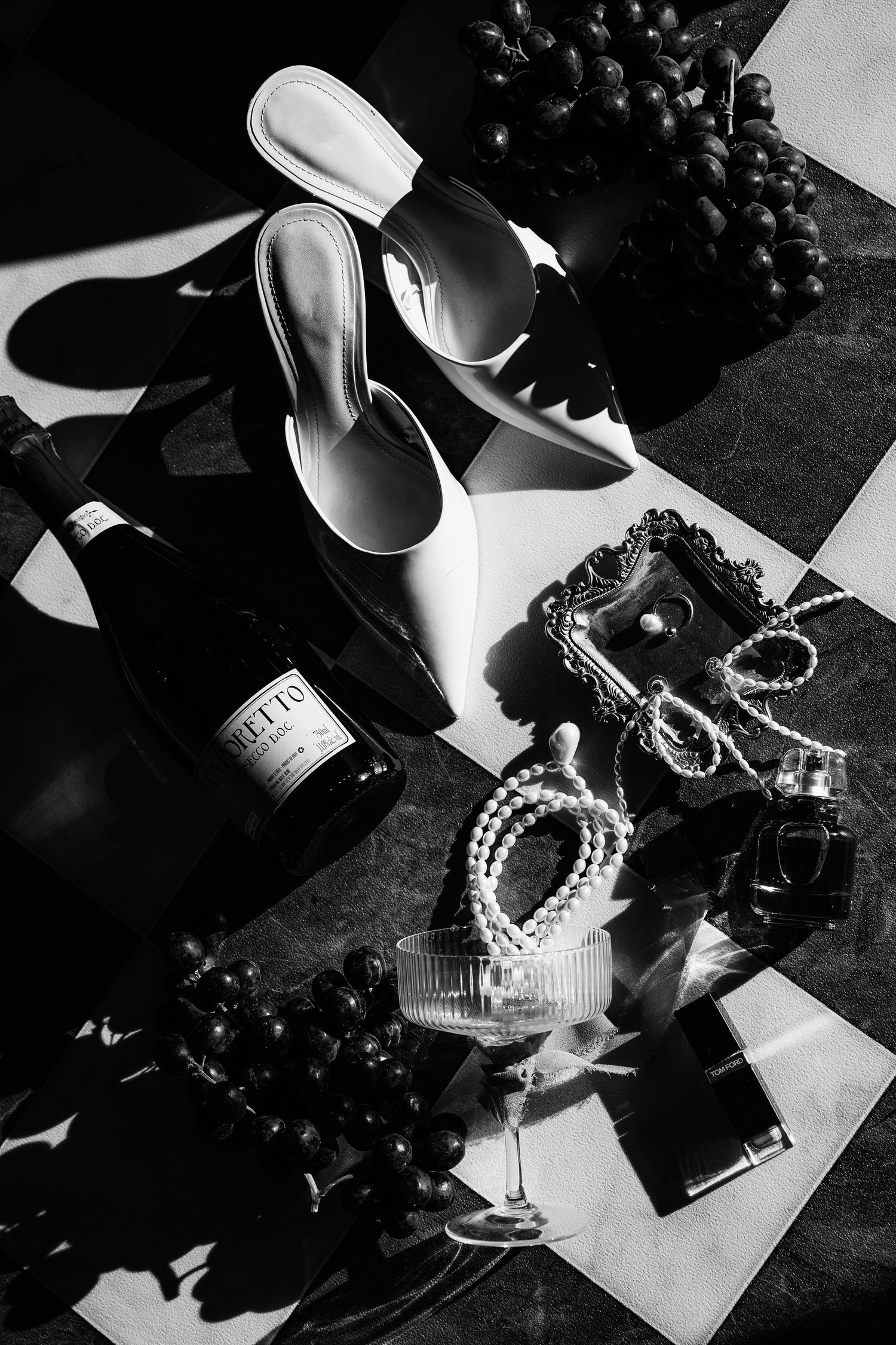 Black and white still life of wedding accessories including pearls, shoes, and champagne glasses.