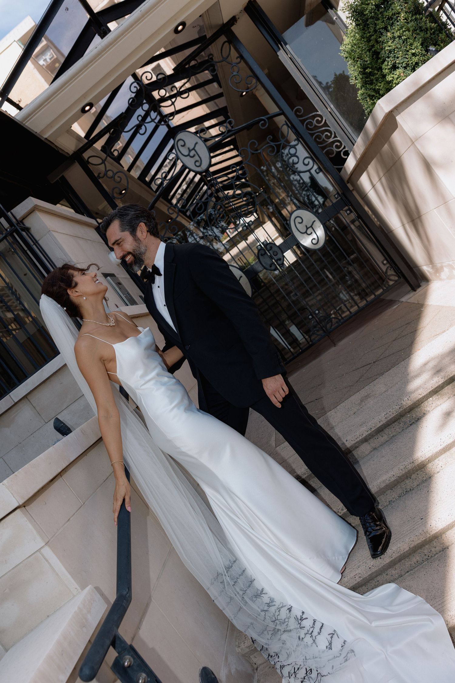 Elegant wedding couple posing on steps near a glass-roofed architectural structure.