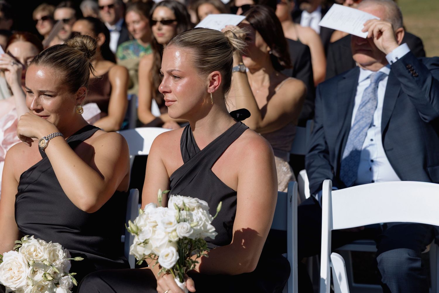 Wedding guests in formal attire seated in white chairs at an outdoor ceremony.