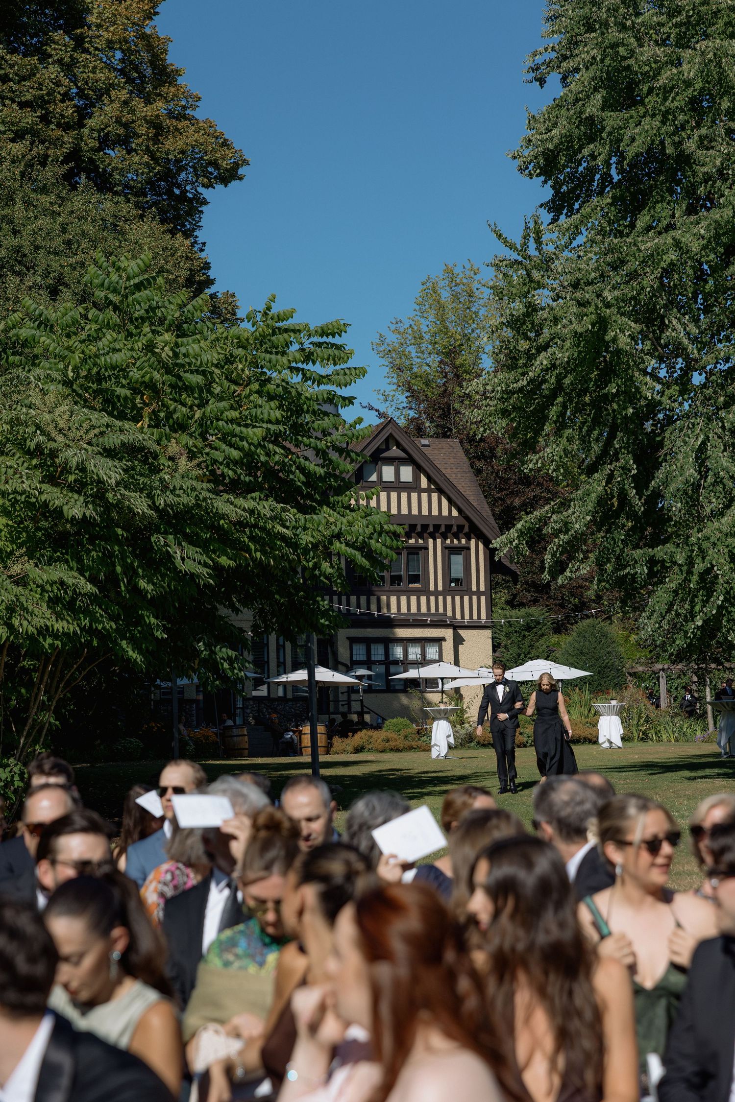 Large crowd gathers for an outdoor wedding ceremony with Tudor-style buildings visible in the background.
