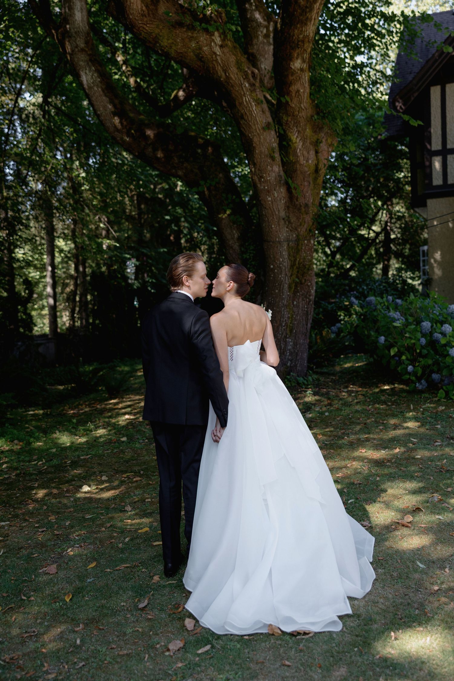 Wedding couple walking together under tree canopy in outdoor garden setting.