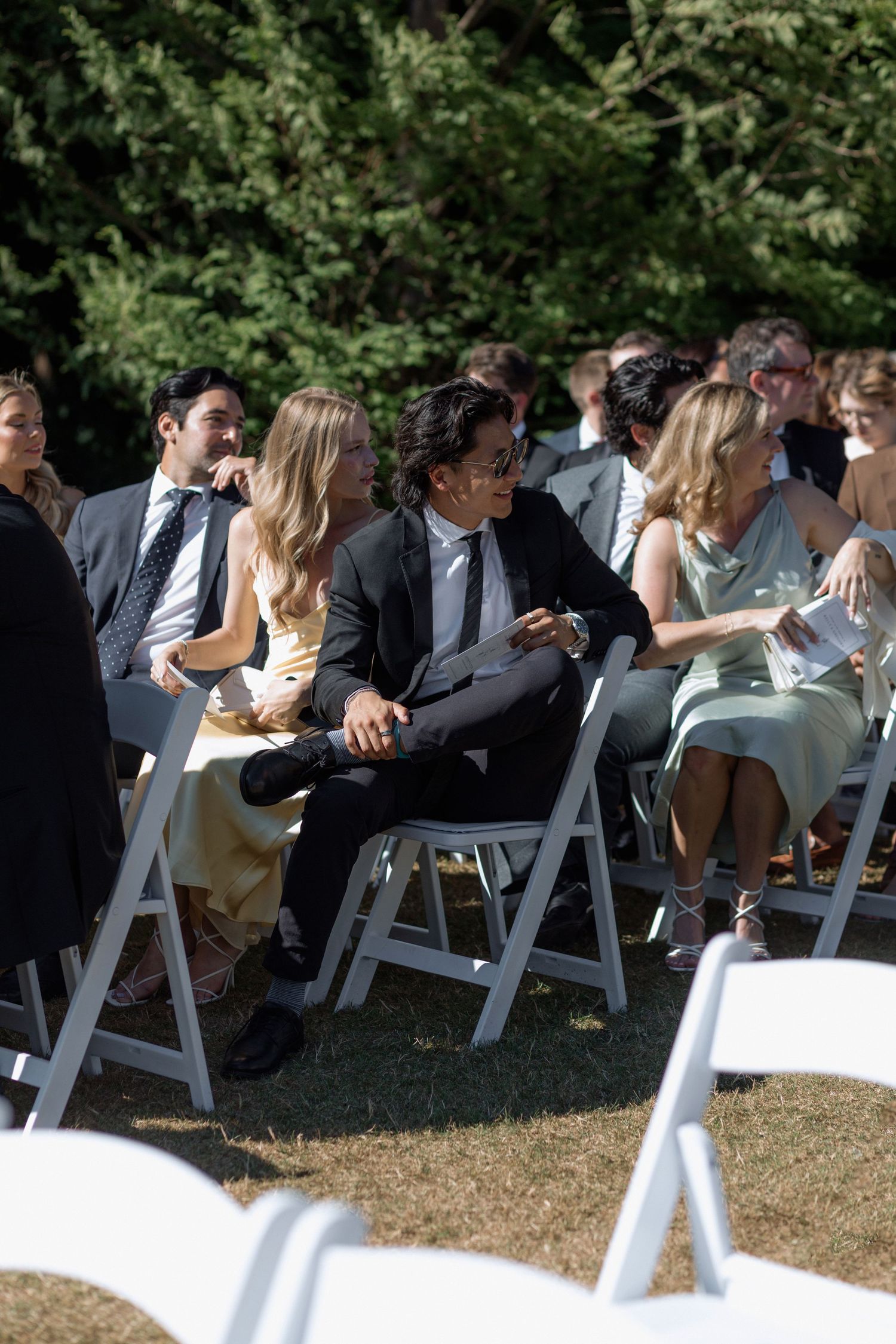 Wedding guests seated in white folding chairs on a sunny lawn during ceremony.