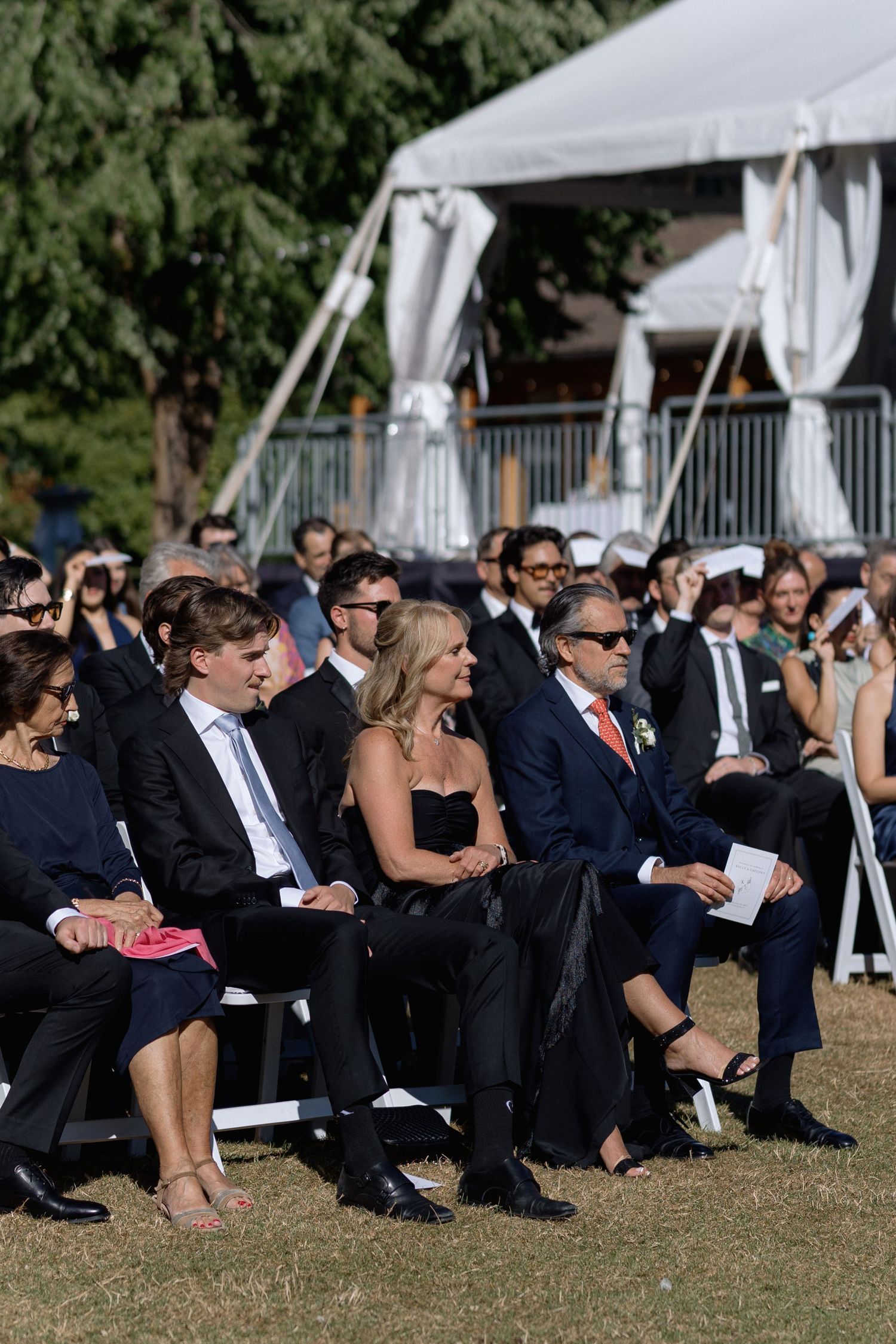 Wedding guests in formal black attire seated on white chairs on a grassy lawn.