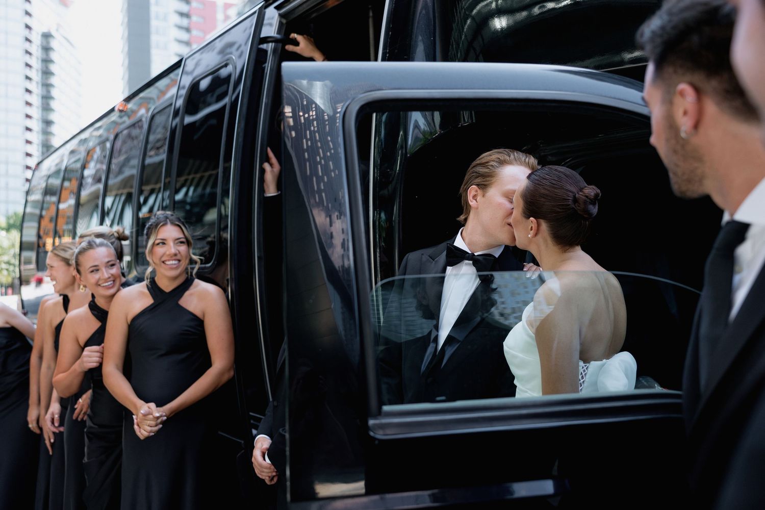 Wedding party members share laughs near black limousine during photo session.