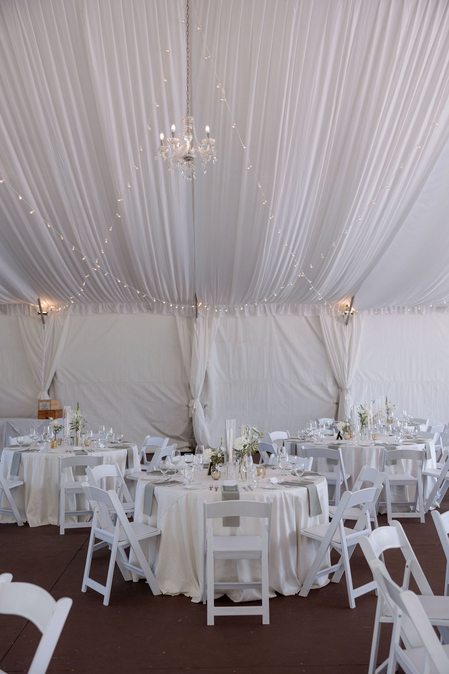 Elegant wedding reception setup with white draped ceiling, crystal chandeliers, and round tables with white linens.
