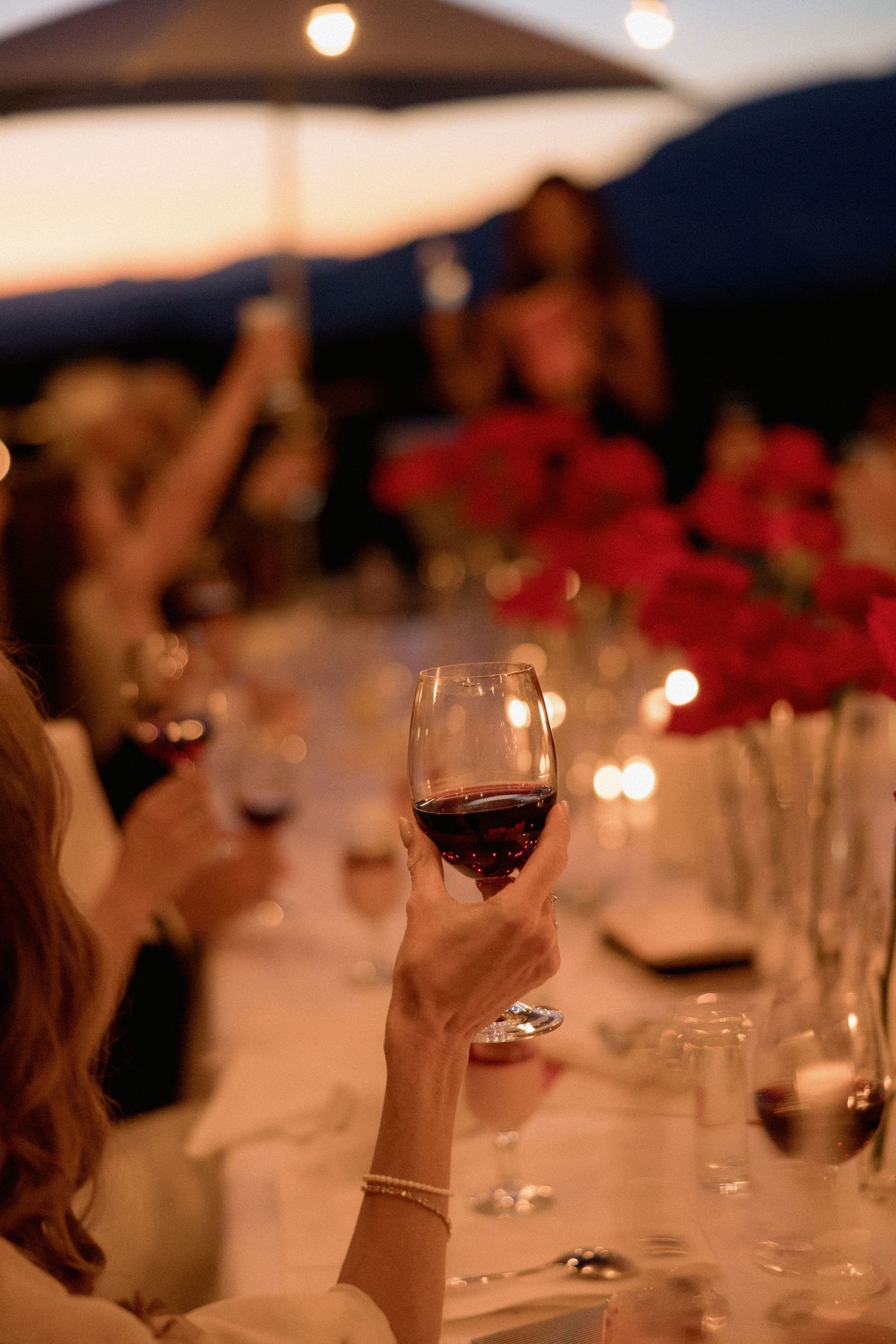 Hand holding a glass of red wine at an elegant outdoor dinner party with bokeh lights.