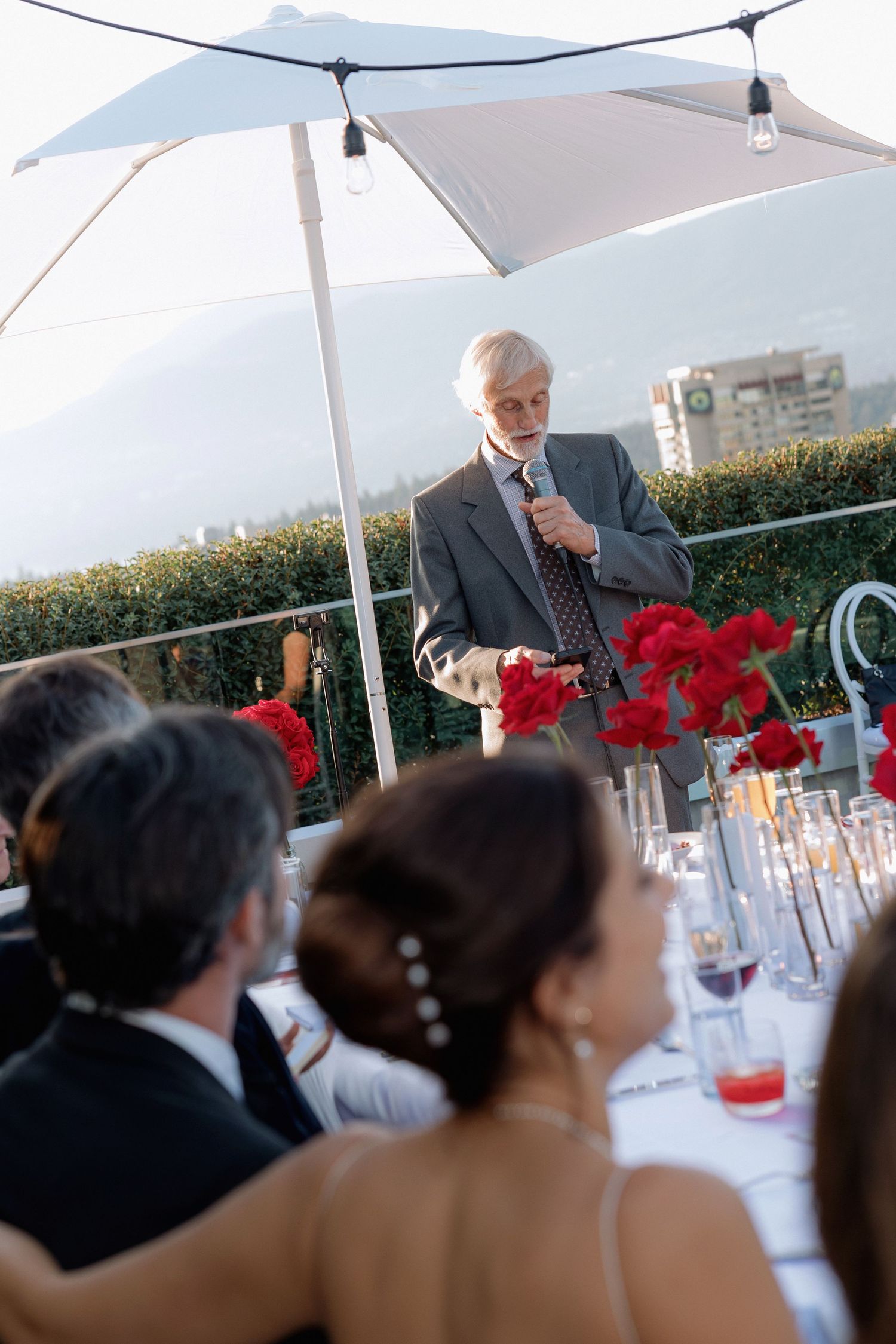 Speaker giving toast at outdoor wedding reception under white canopy with red floral decor.
