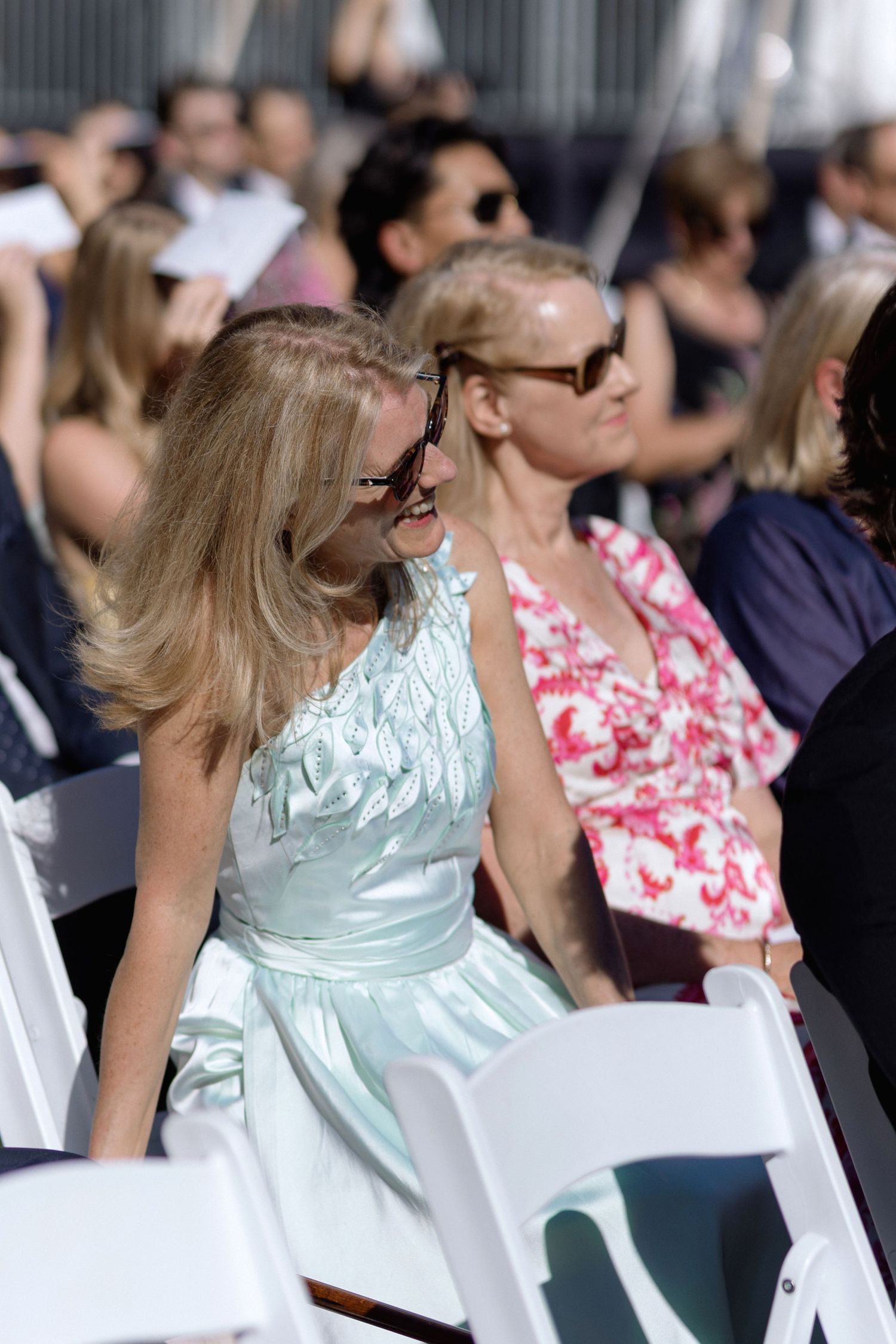 Wedding guests in colorful dresses and sunglasses seated on white chairs outdoors.