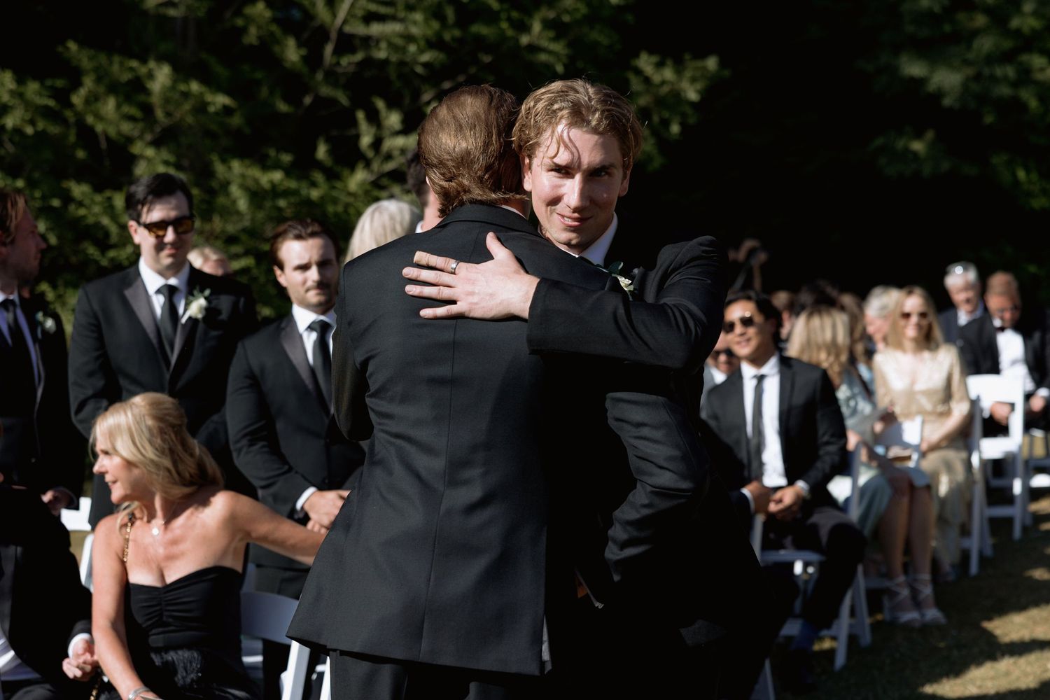 Guests in formal attire socialize at an outdoor wedding venue with white chairs and natural surroundings.