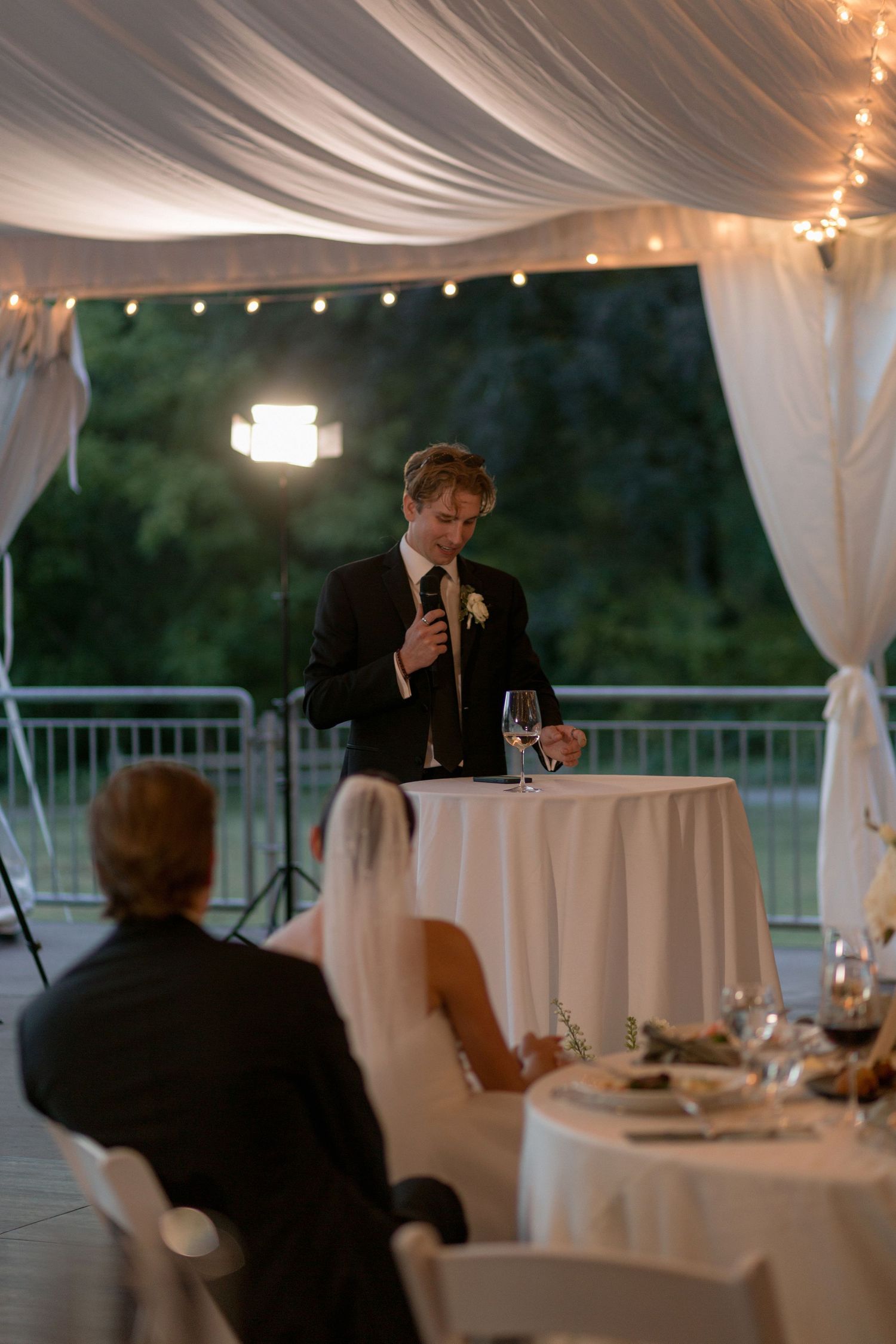 Evening wedding speech being delivered in an elegant tent-covered venue with string lights.