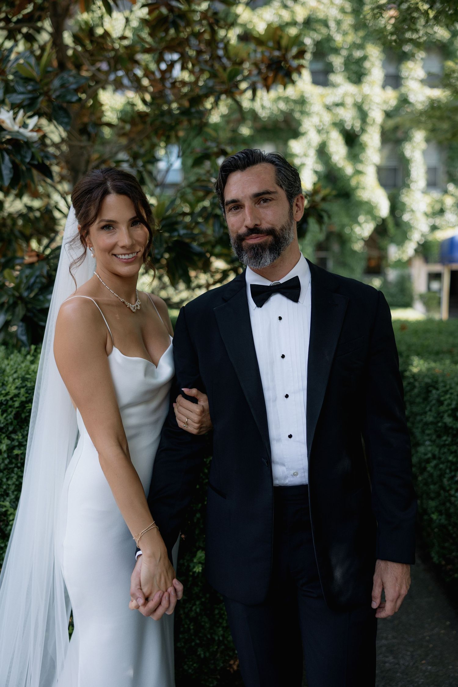 A bride and groom share a tender moment outdoors with lush greenery in the background in front of the Sylvia Hotel in Vancouver