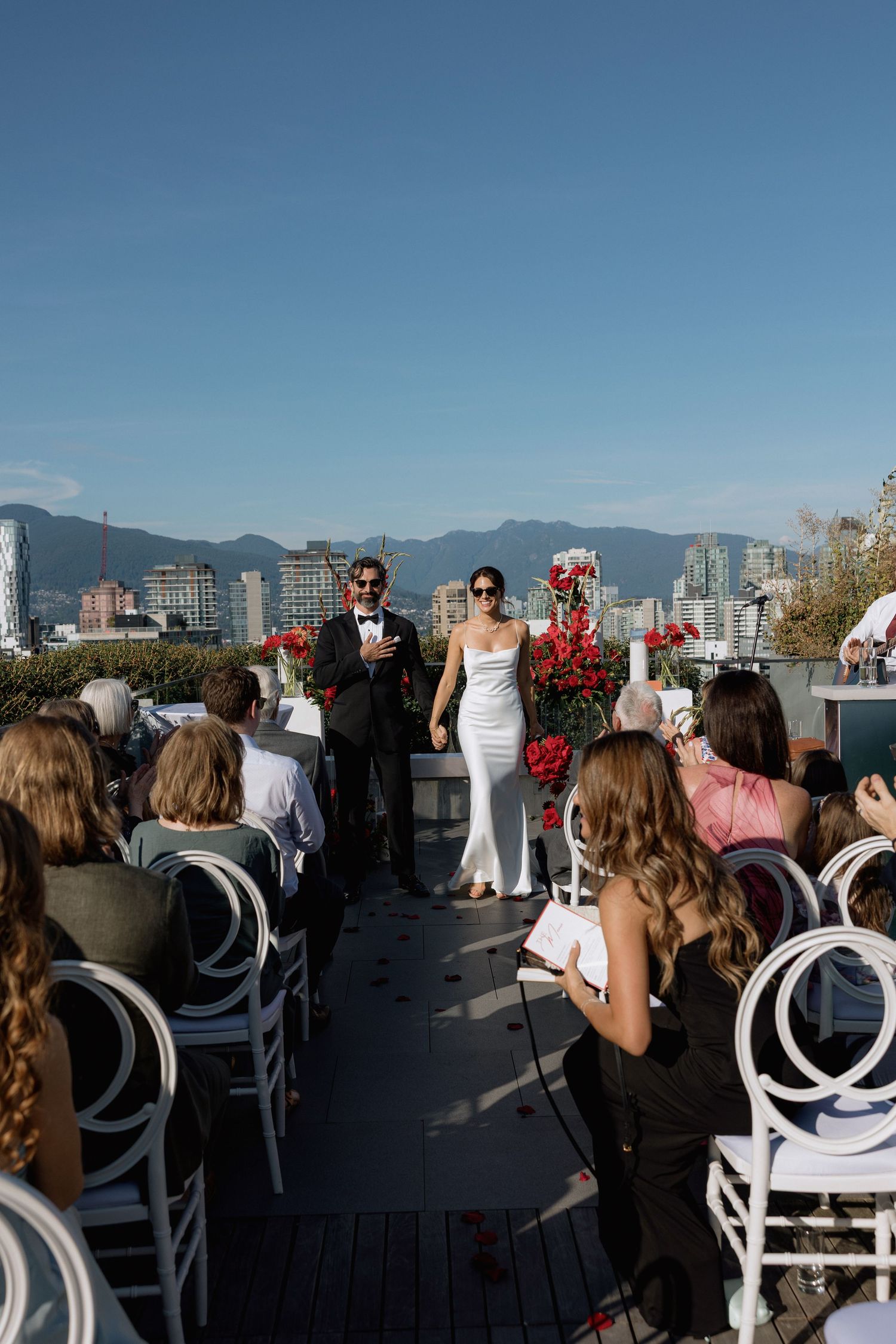 Rooftop wedding ceremony with city skyline and mountains in background featuring white chairs and red floral decorations.