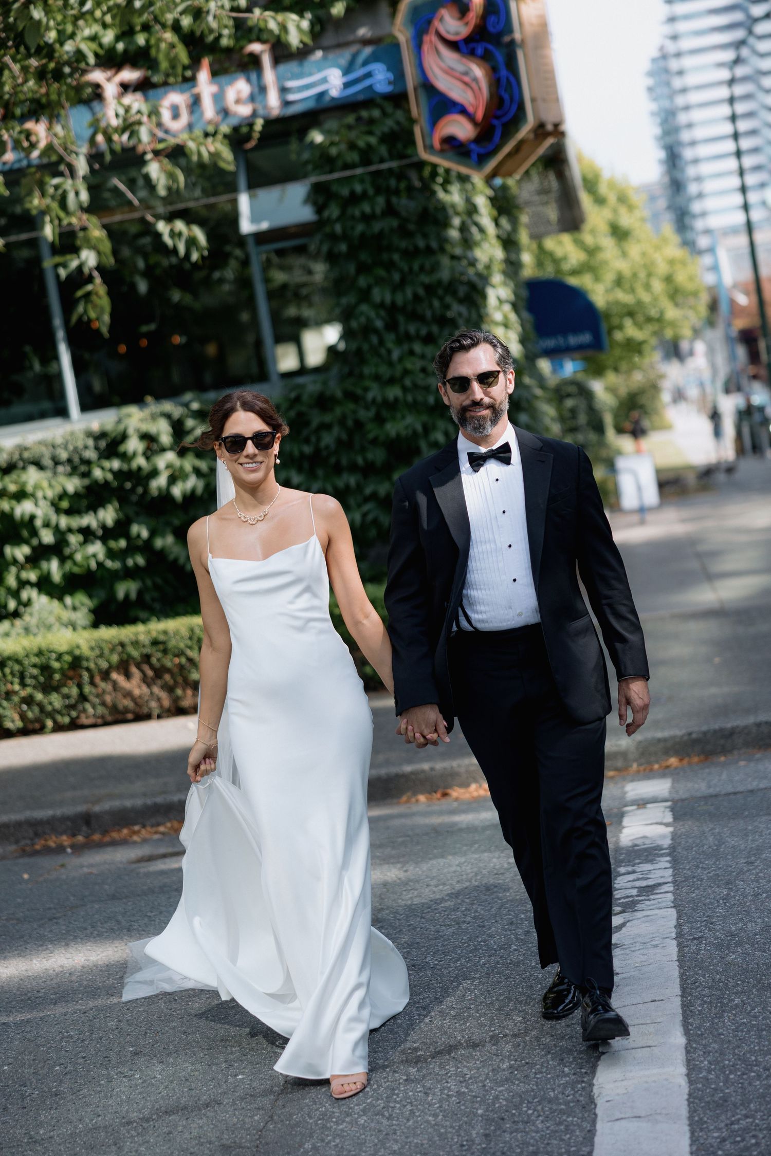 A couple in wedding attire walks together on a city street with trees and buildings in the background in front of the Sylvia Hotel