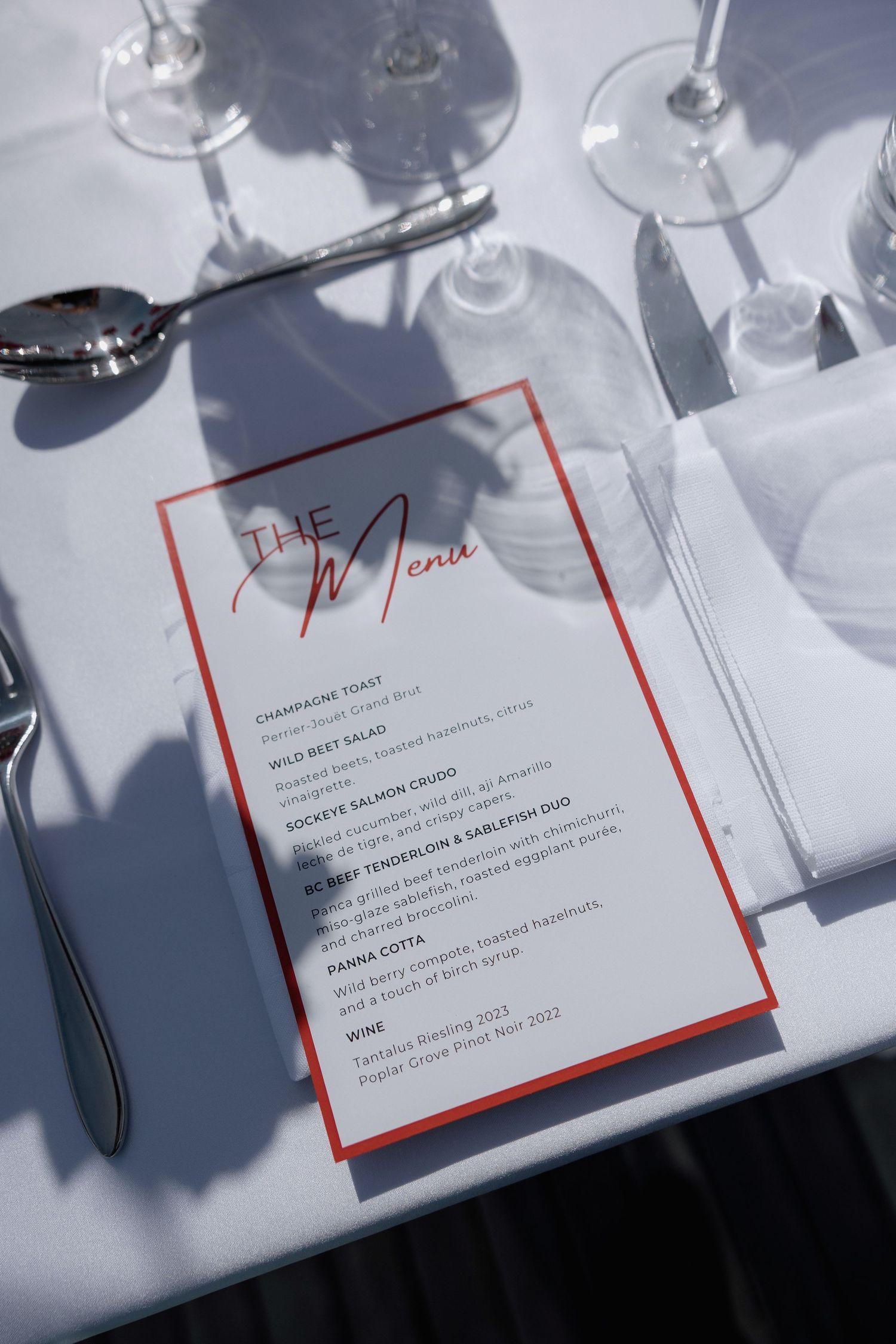 Elegant restaurant menu cards with red borders displayed on a white table with wine glasses casting shadows.