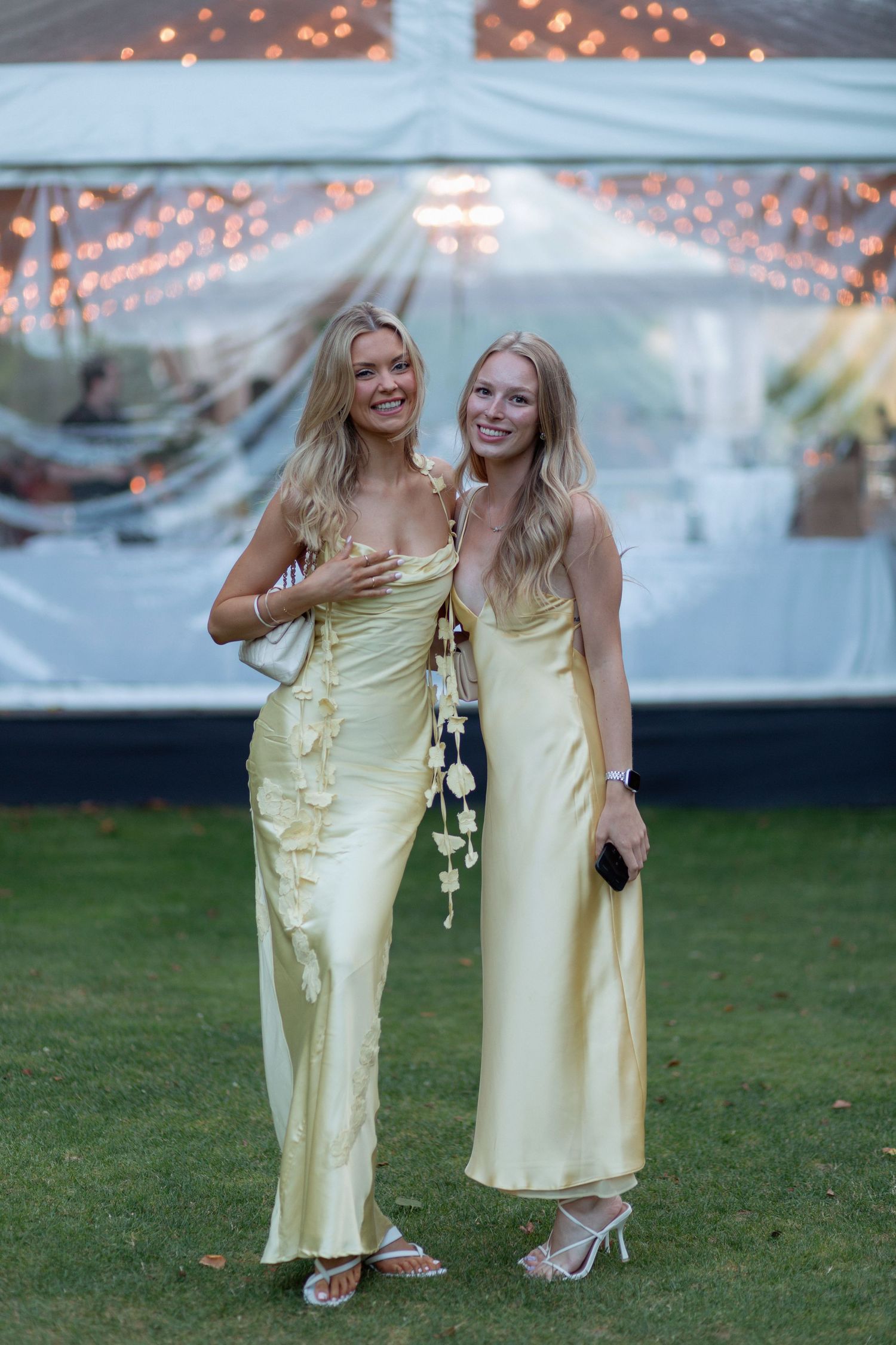 Two bridesmaids in pale yellow dresses pose together under string lights at an outdoor reception.
