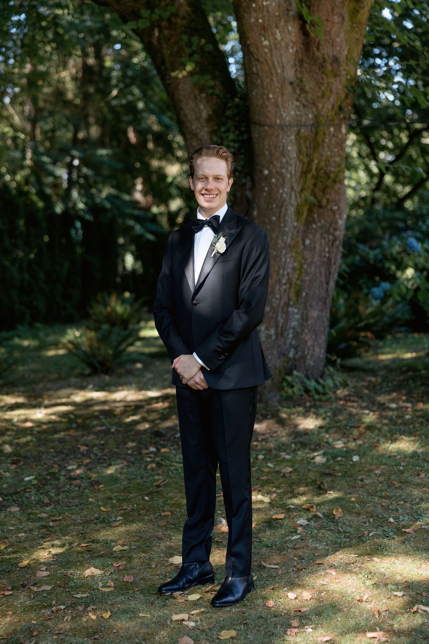 Ring bearer in classic black tuxedo poses against tree-lined backdrop at outdoor wedding.