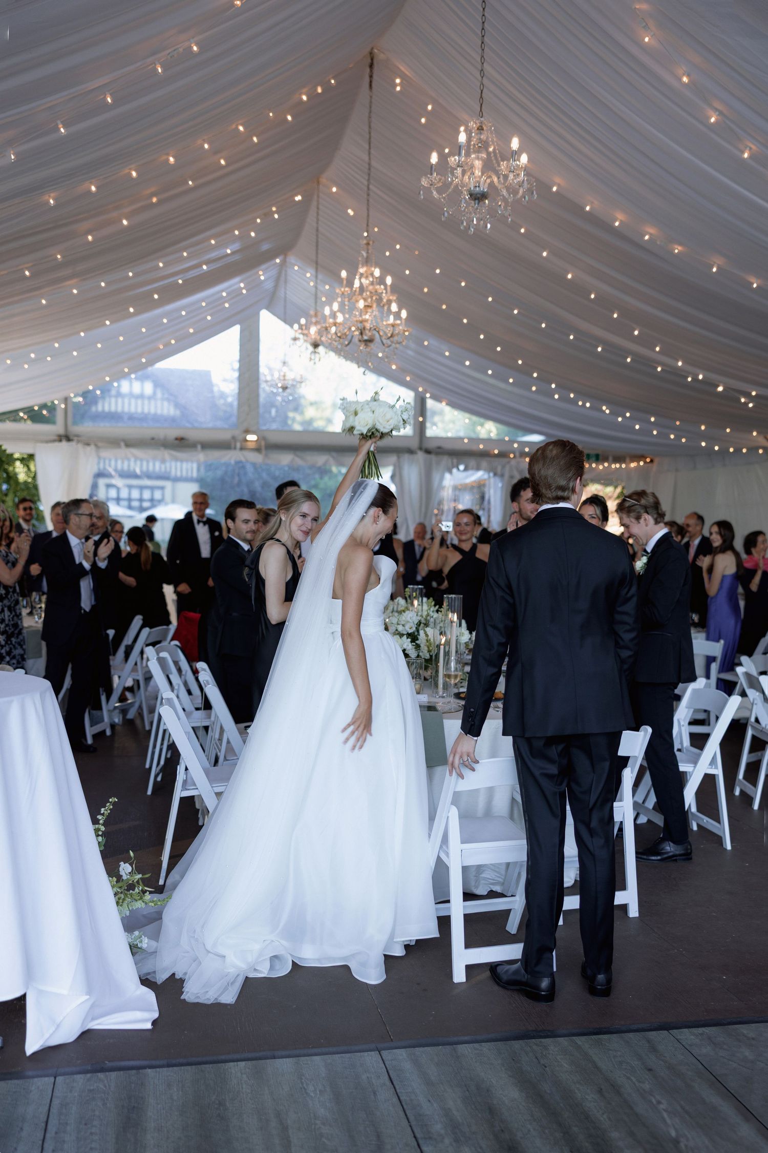 Wedding reception in progress inside elegant white tent with chandeliers and string lights.