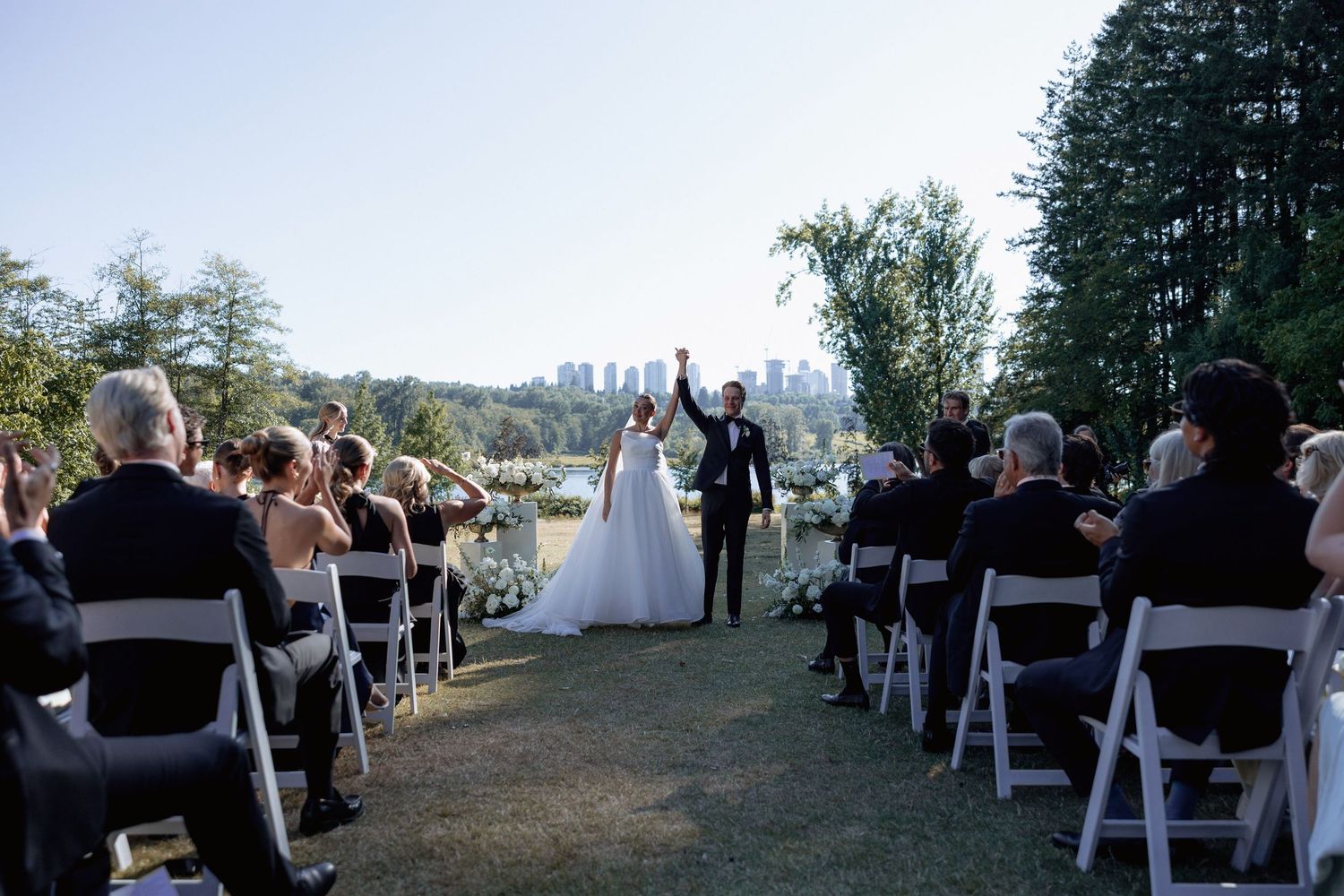 Wedding guests seated in rows of white chairs at an outdoor ceremony with a cityscape backdrop and Deer Lake