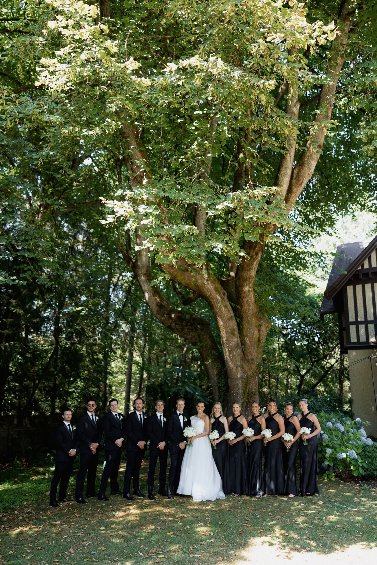 Wedding party lines up under tall trees in a wooded outdoor setting.