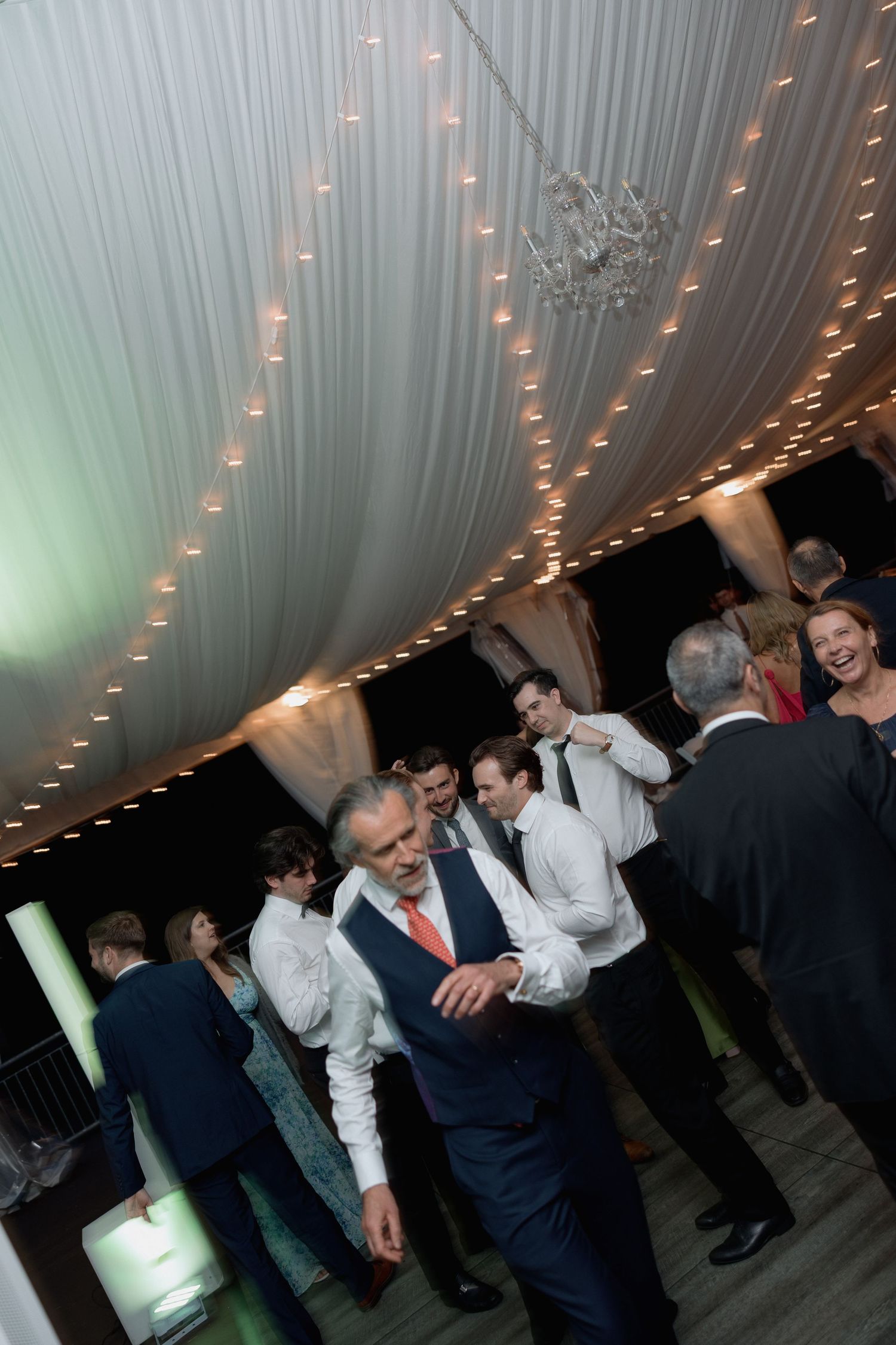 Reception guests dancing beneath elegant draped ceiling with twinkling lights and chandelier.