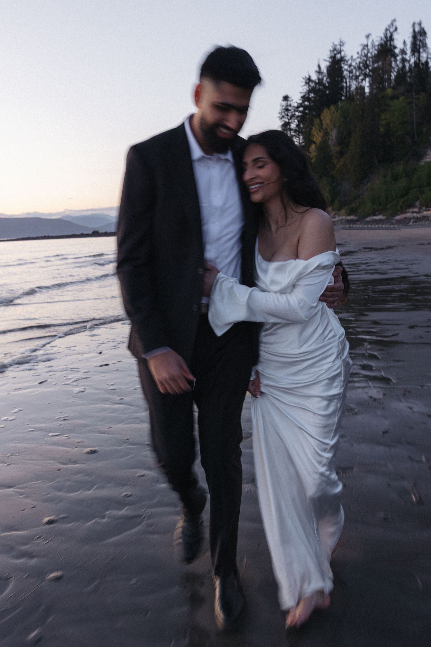 A couple in formal attire embraces on a beach at sunset with evergreen trees in the background.