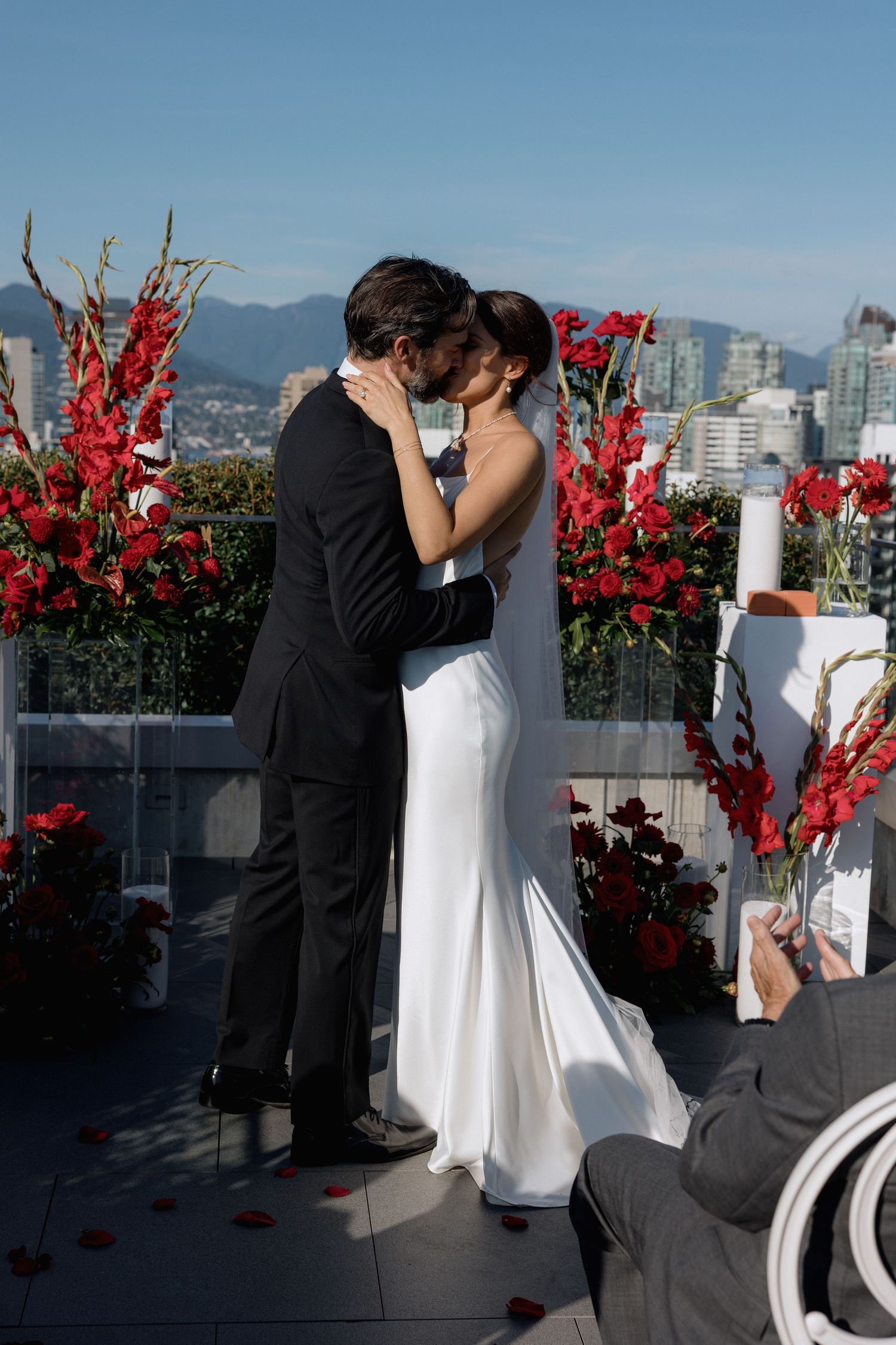 Couple sharing intimate moment during rooftop ceremony with red flowers and city views.