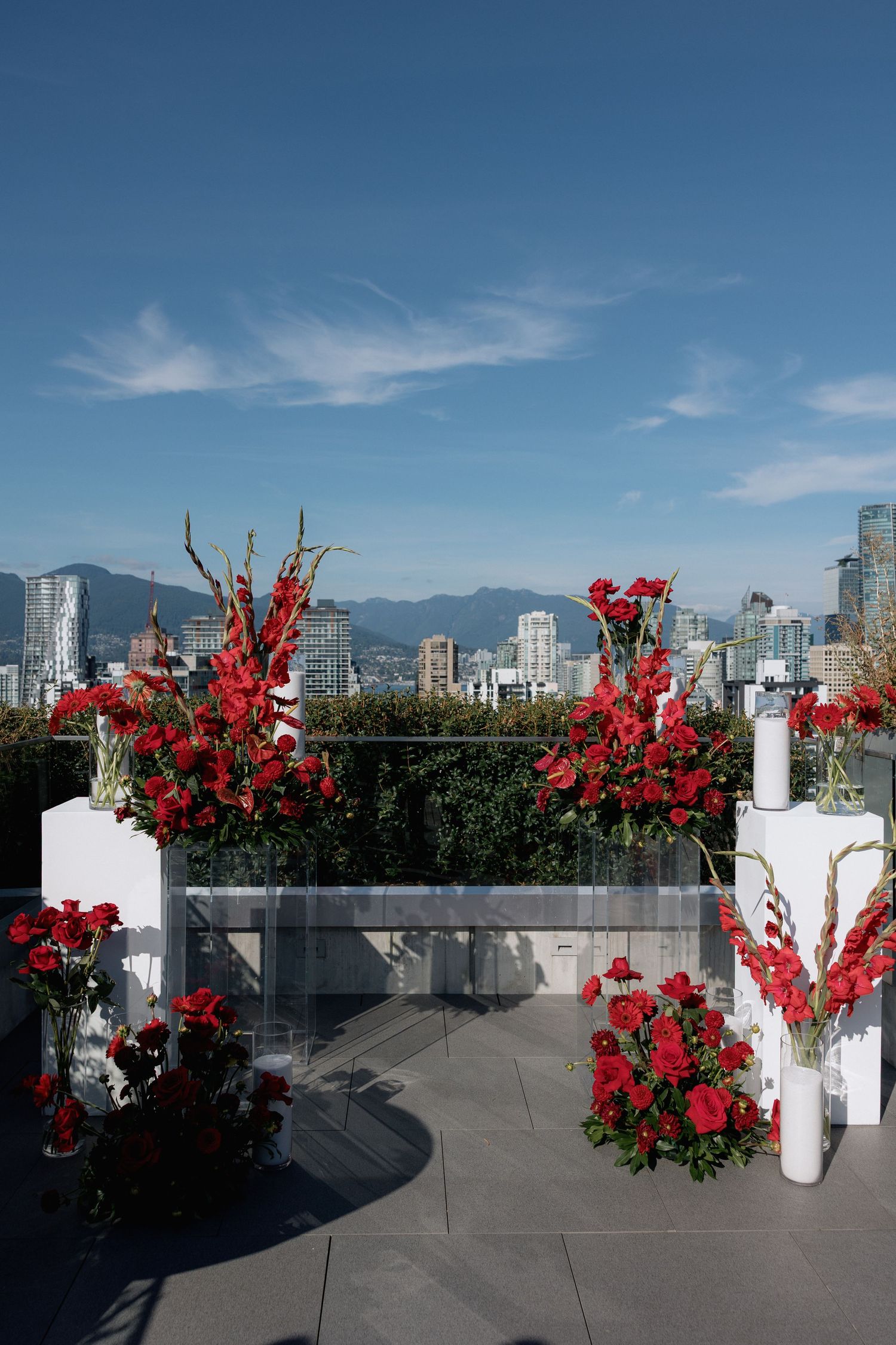Red floral arrangements on white pedestals decorate a rooftop terrace with Vancouver's skyline and mountains in the background.