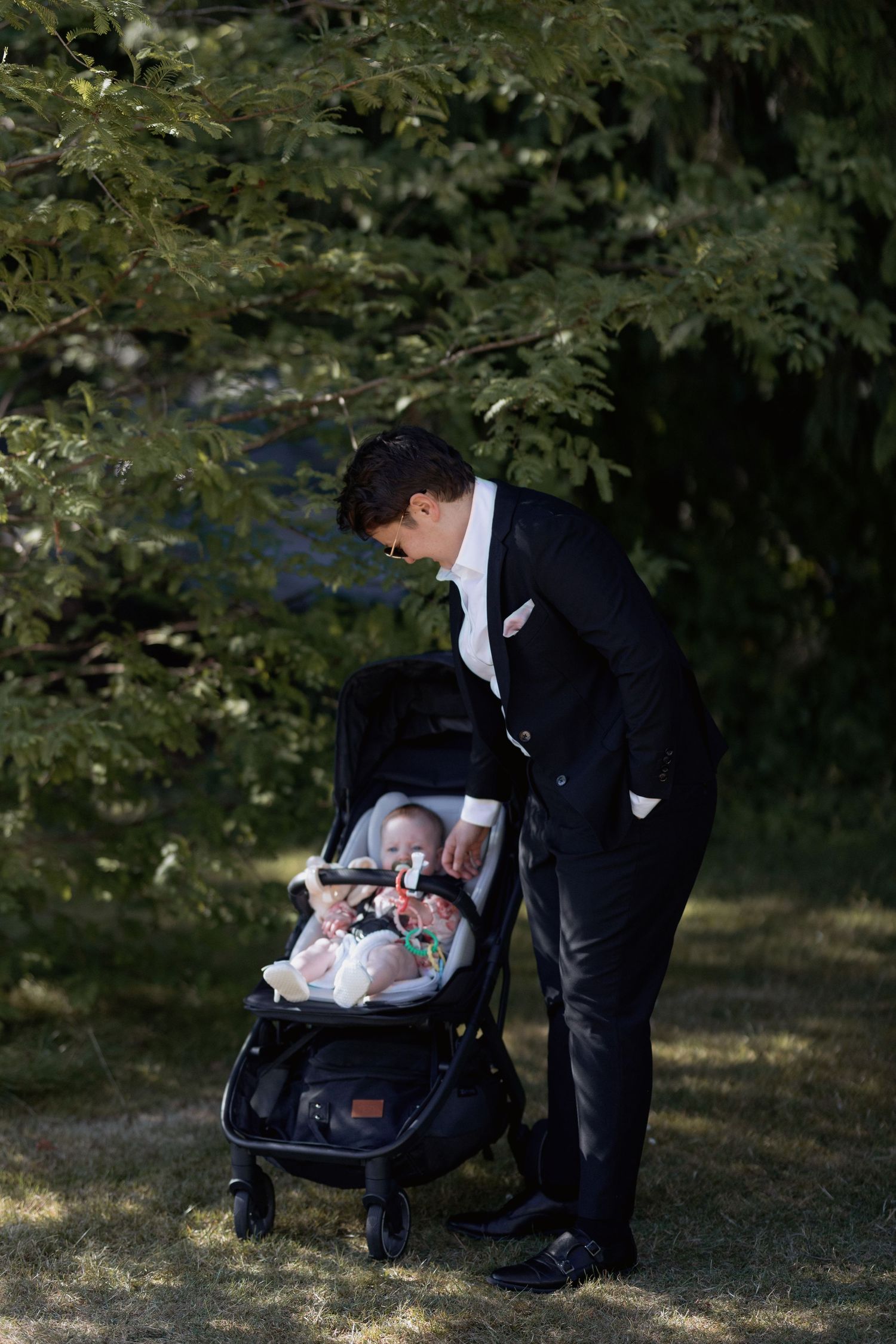 Person in formal suit tending to baby in stroller under shaded trees at wedding.