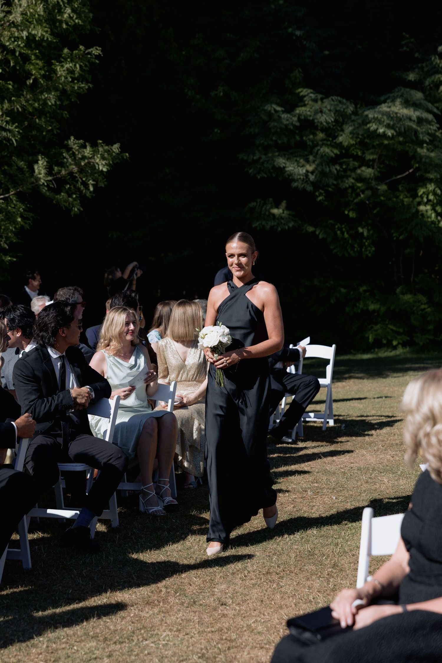 Guests mingle before an outdoor wedding ceremony on a sunny afternoon in a wooded setting.