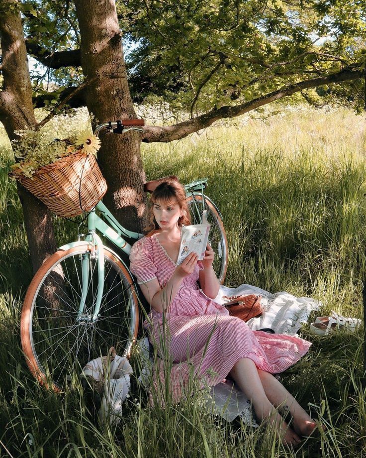 Woman in pink dress reading book beside vintage green bicycle under tree in sunny meadow.