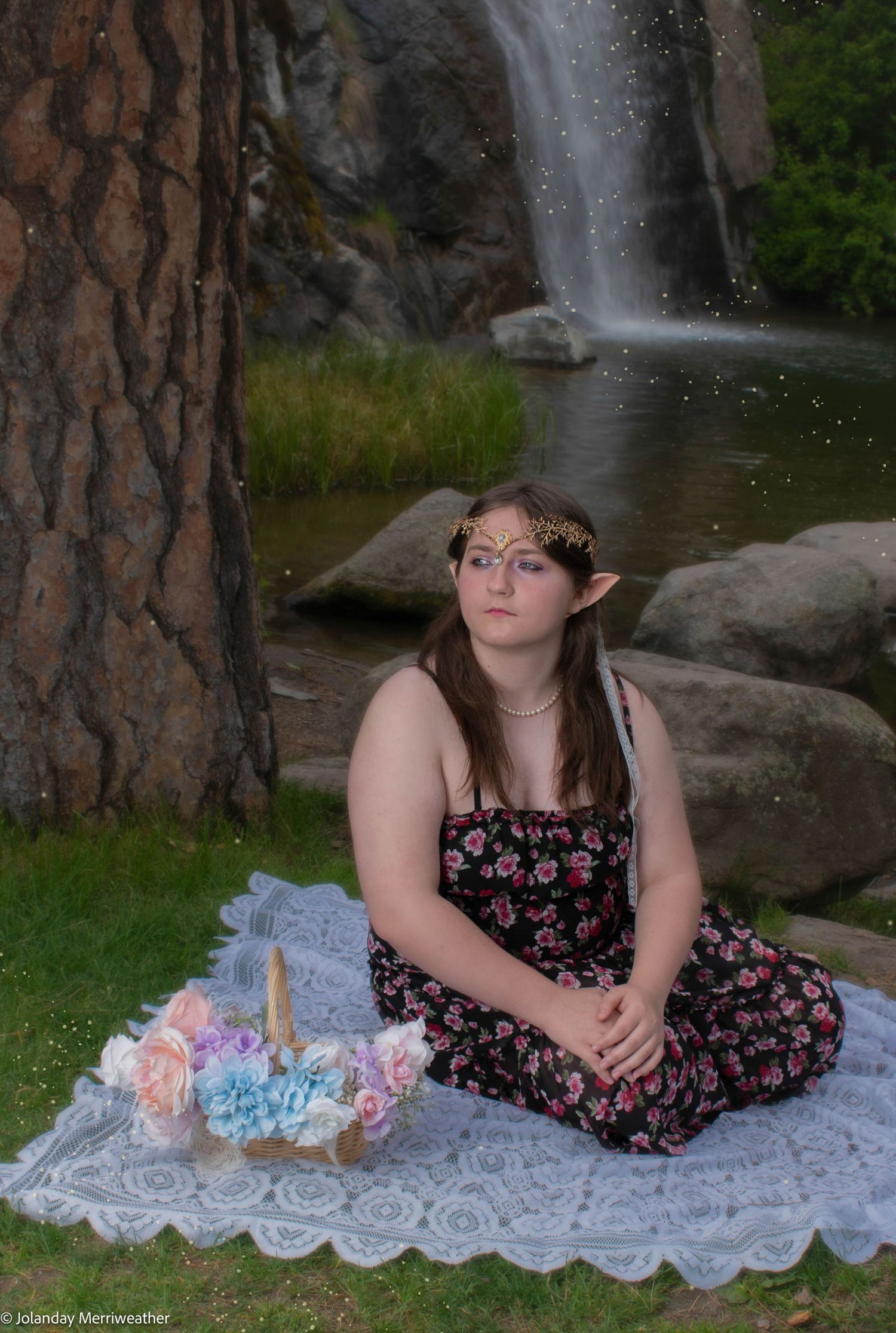 Young woman in elf costume sitting by waterfall with floral arrangement in forest.