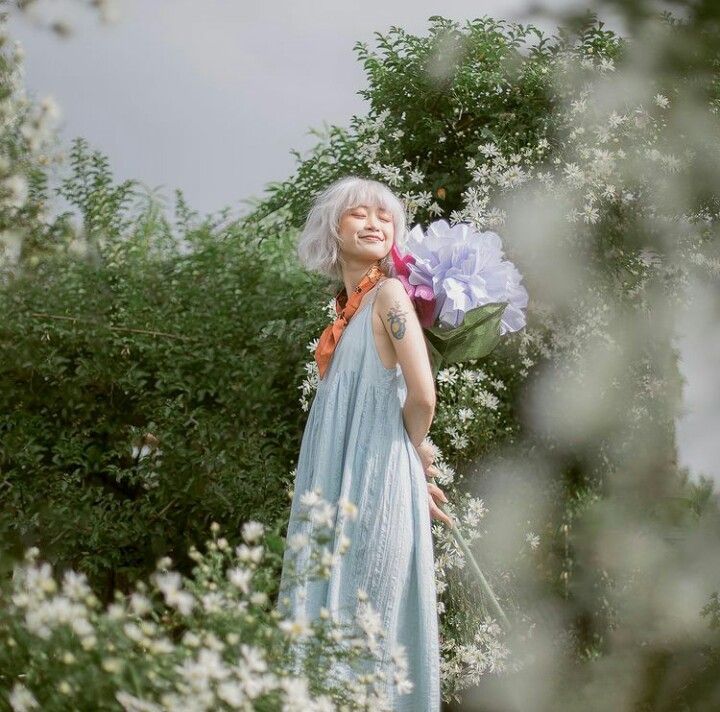 Smiling woman in light blue dress with oversized purple flower among white wildflowers outdoors.