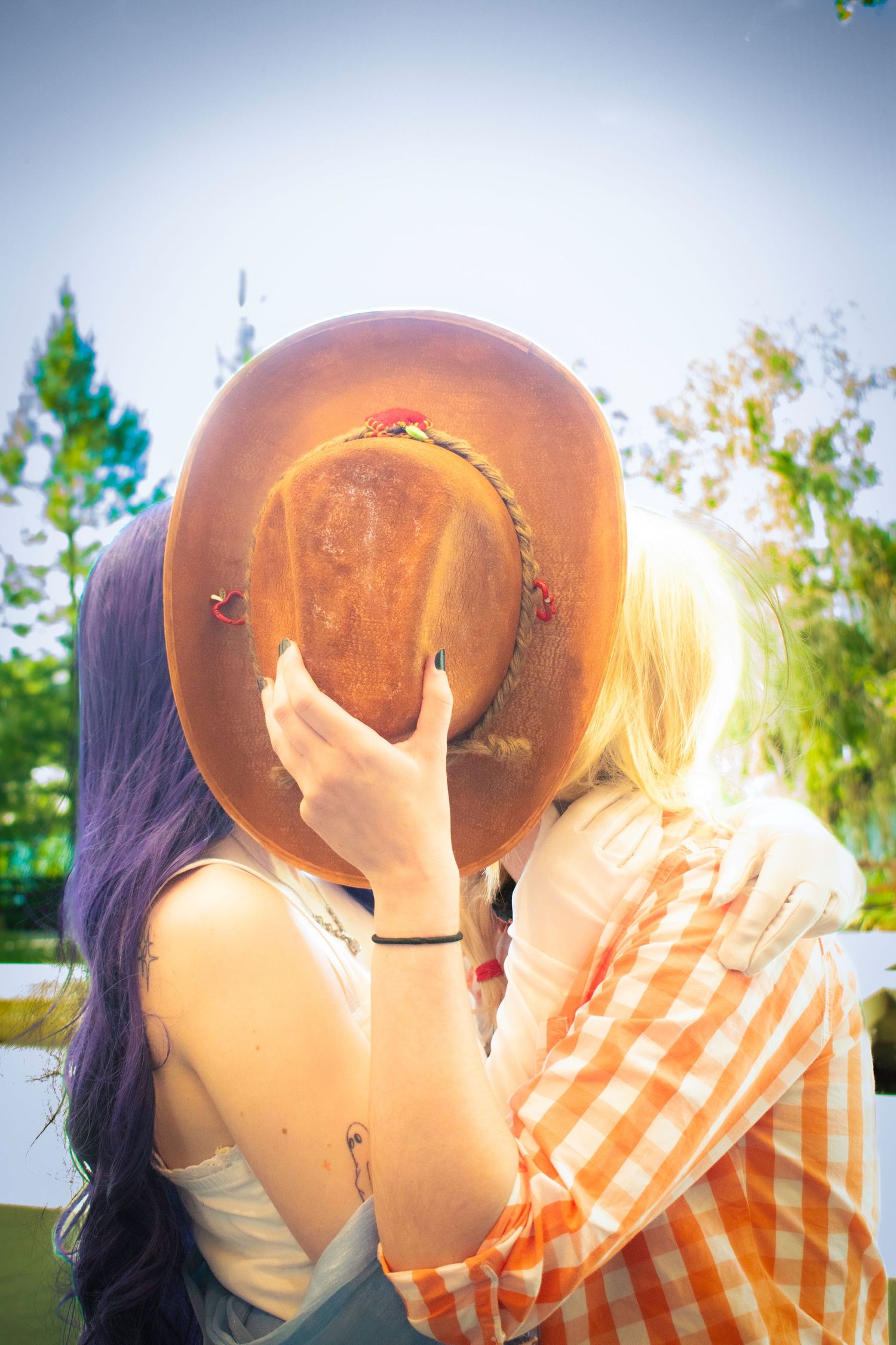 Two people sharing a kiss hidden behind a brown cowboy hat outdoors, one with purple hair, one blonde.
