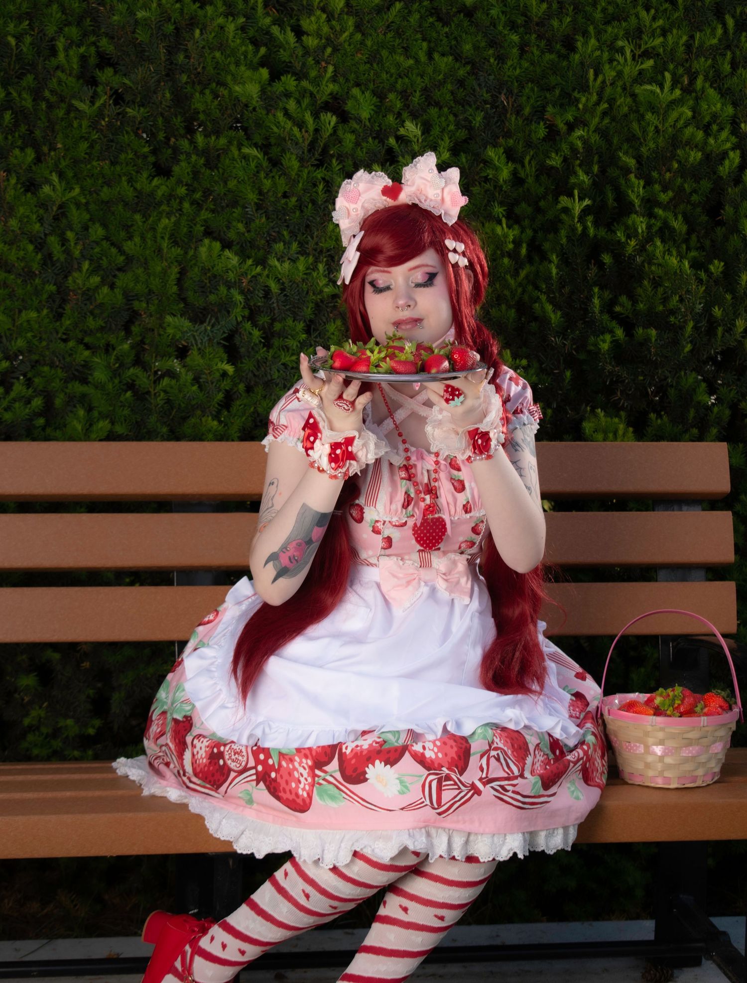 A woman in a strawberry-themed Lolita dress with red wig sits on a bench holding strawberries, wearing striped tights and red shoes.