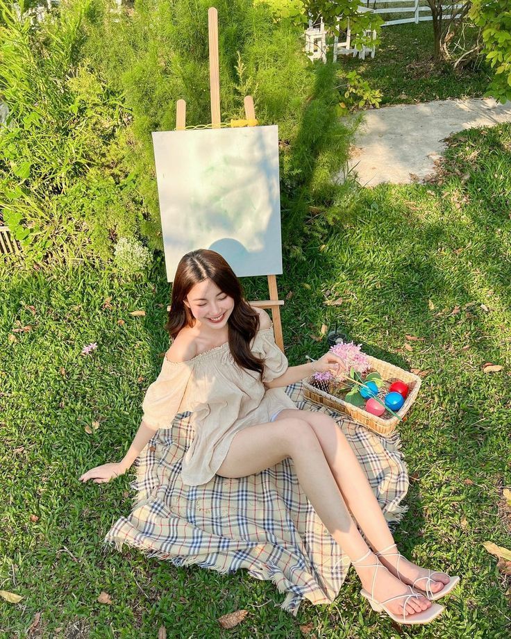 Smiling woman in beige outfit sitting on plaid blanket beside blank canvas easel in garden.