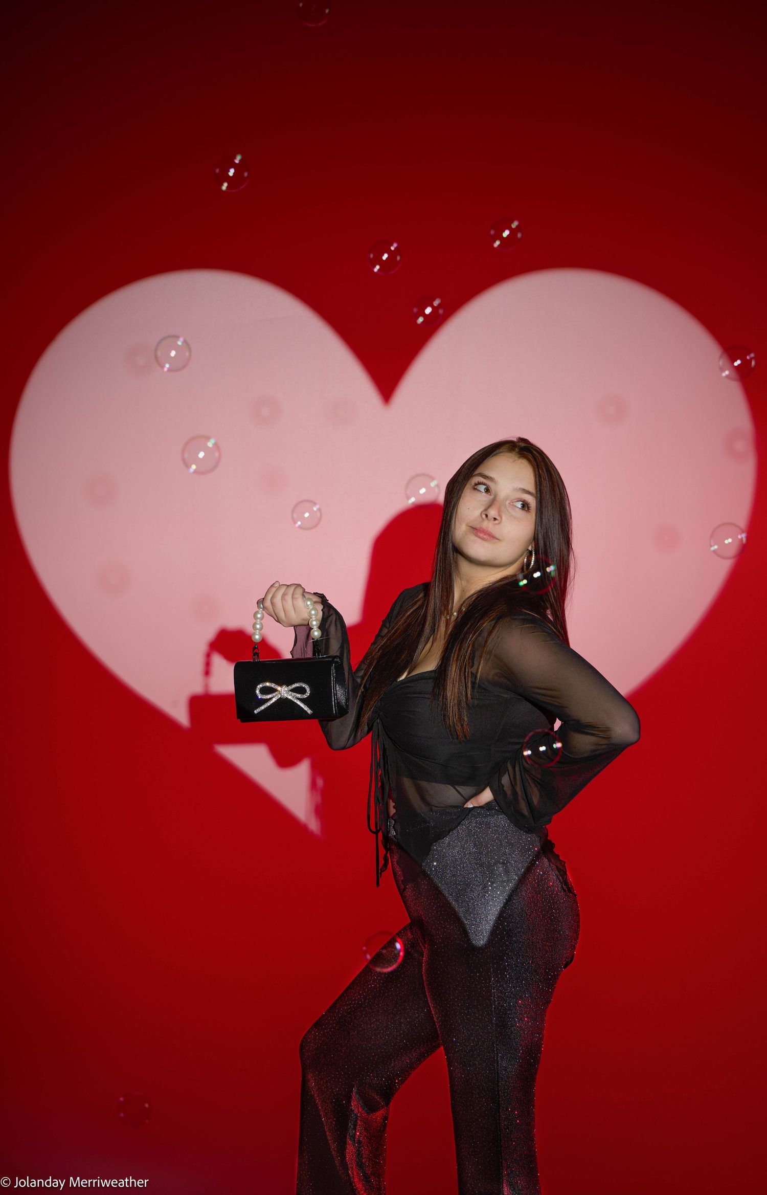 Young woman in black outfit posing in front of a red heart backdrop with floating bubbles.