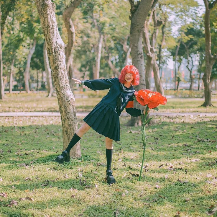 Girl in dark school uniform with pink hair posing beside giant red flower in park.