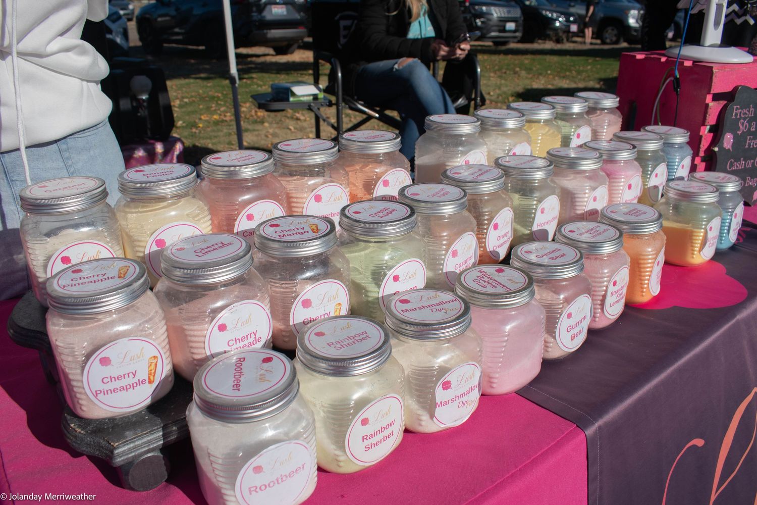 Colorful mason jar candles with labels arranged on a table at an outdoor craft fair.