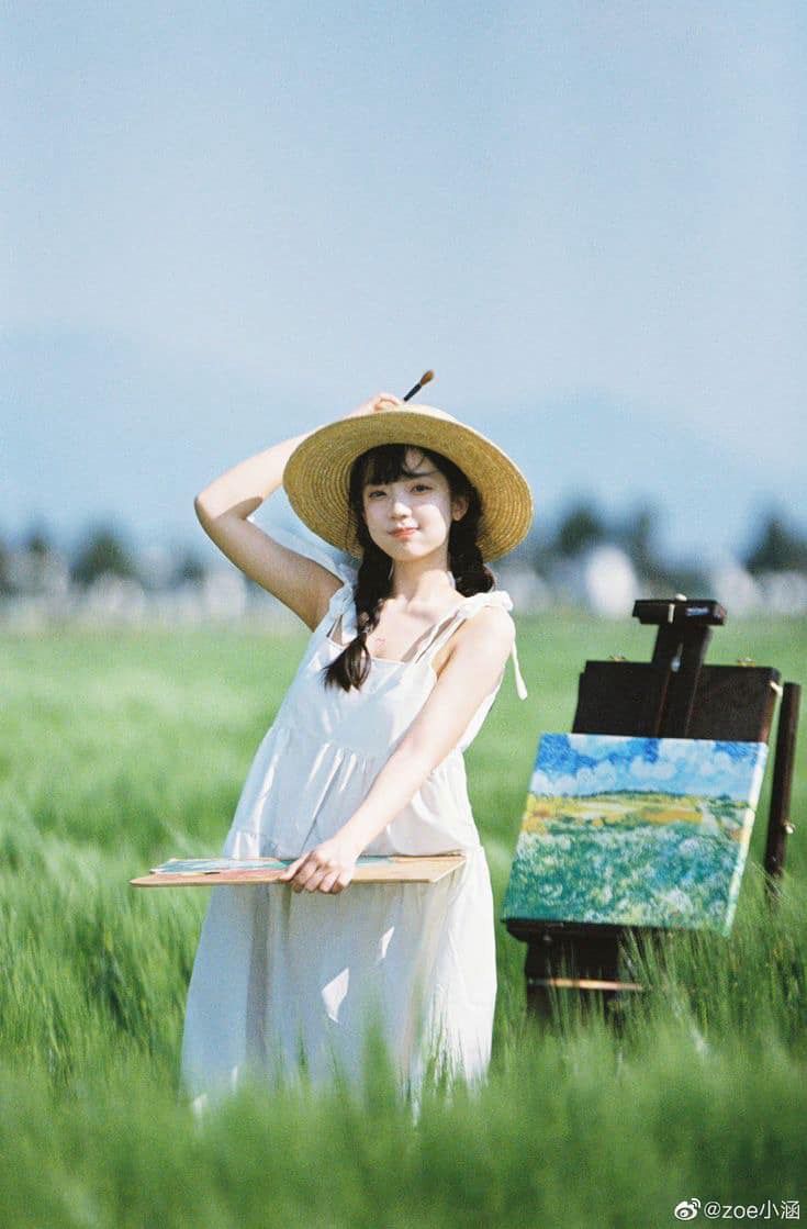 Young woman in white dress and straw hat posing beside painting easel in green wheat field.