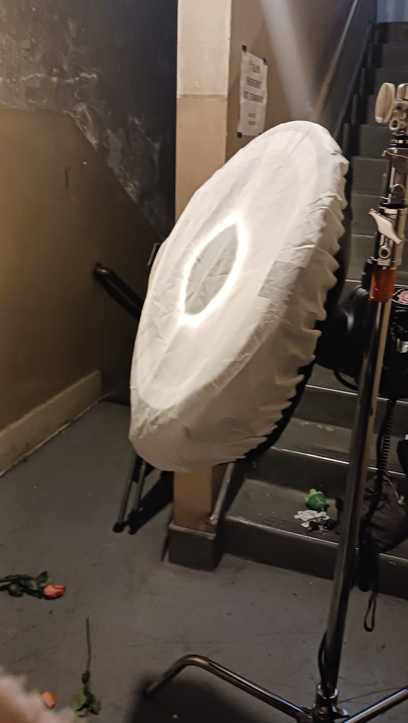 A ring light on a stand illuminates a dark stairwell storage area with equipment.
